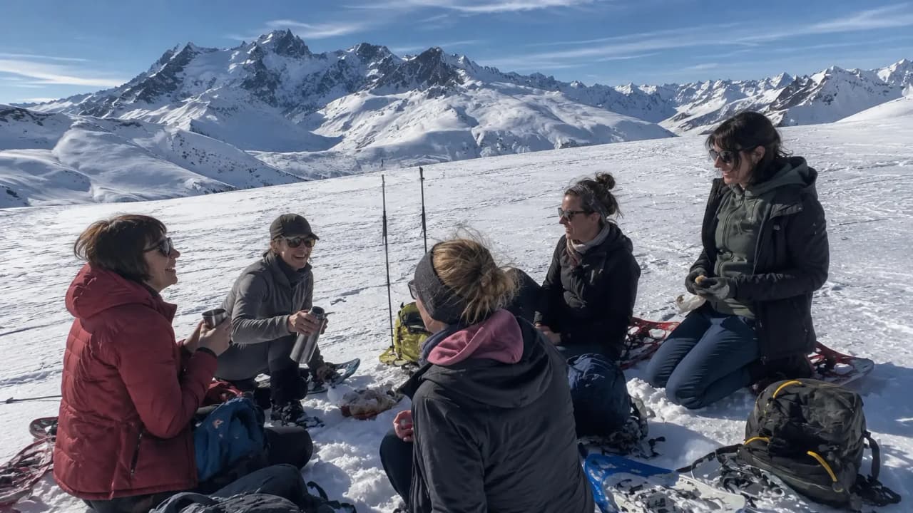 Six women are enjoying a relaxing break, snowshoeing in the sunshine, with a view of the snow-capped mountains.