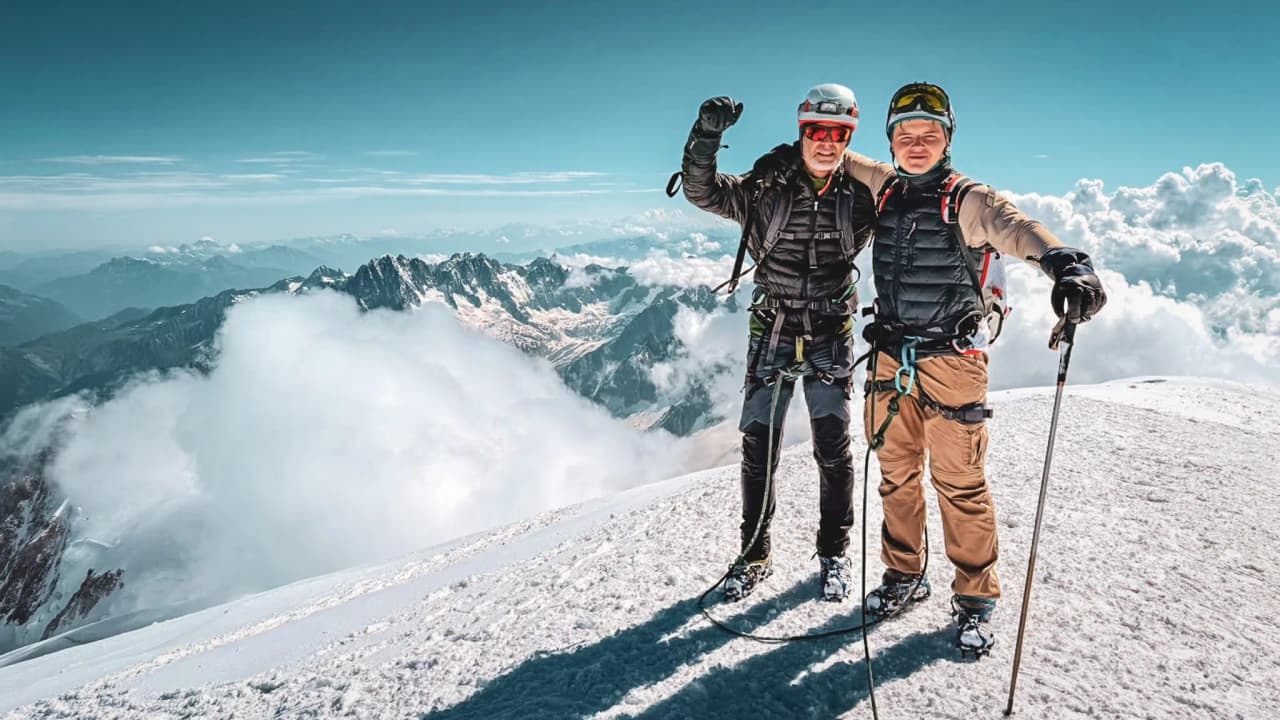 Two smiling mountaineers on the summit of Mont Blanc, surrounded by clouds and majestic mountains.