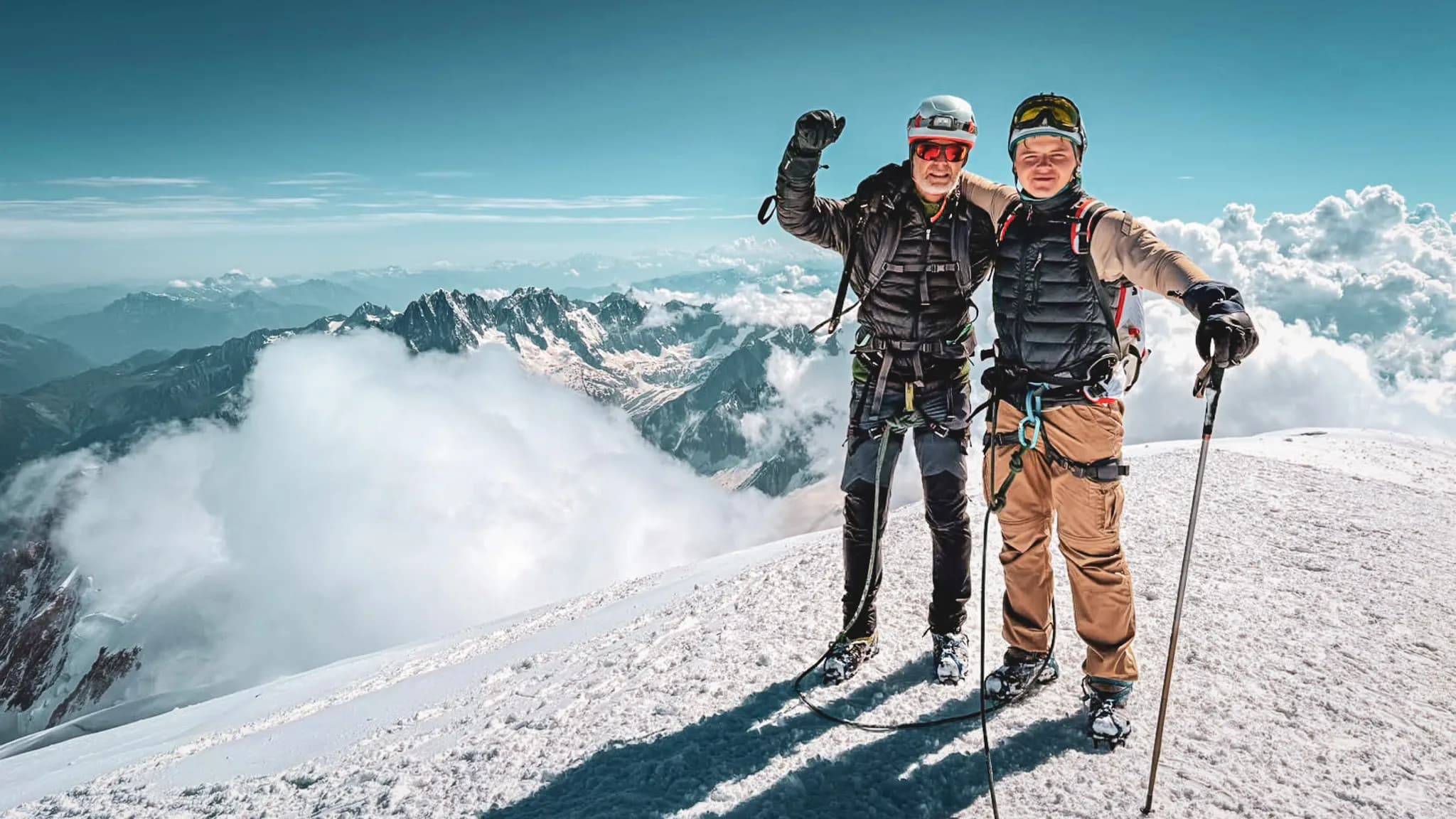 Deux alpinistes souriants au sommet du Mont Blanc, entourés de nuages et de montagnes majestueuses.
