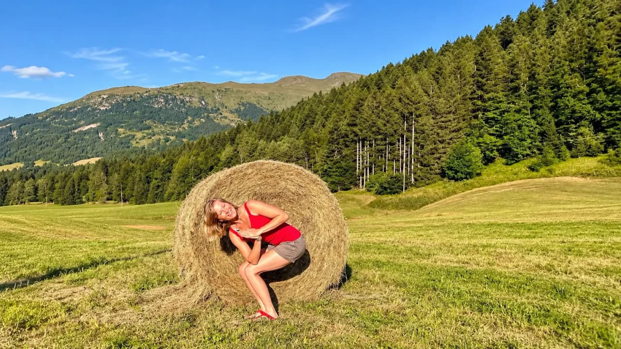 Femme souriante posant près d'une meule de foin, entourée de verdure et de montagnes ensoleillées.