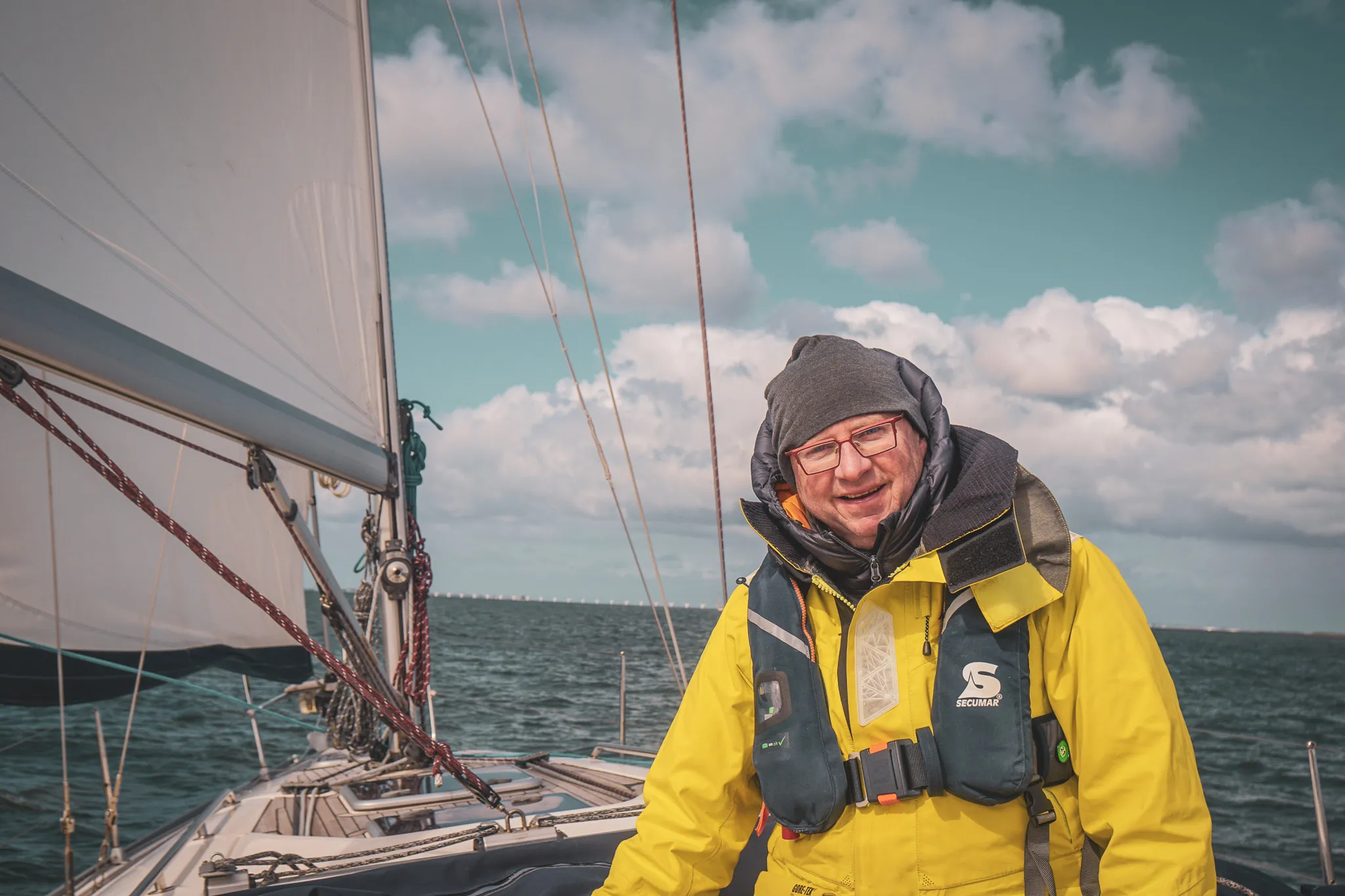 Homme souriant en combinaison jaune sur un voilier, naviguant sur des eaux paisibles sous un ciel nuageux.