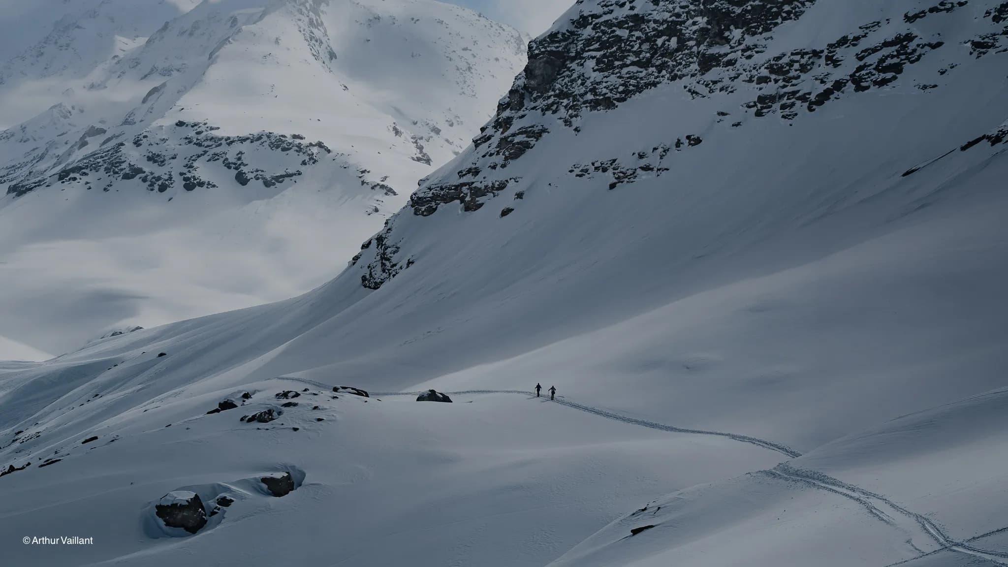 Two skiers are making their way across a vast snowfield, surrounded by majestic Alpine mountains.