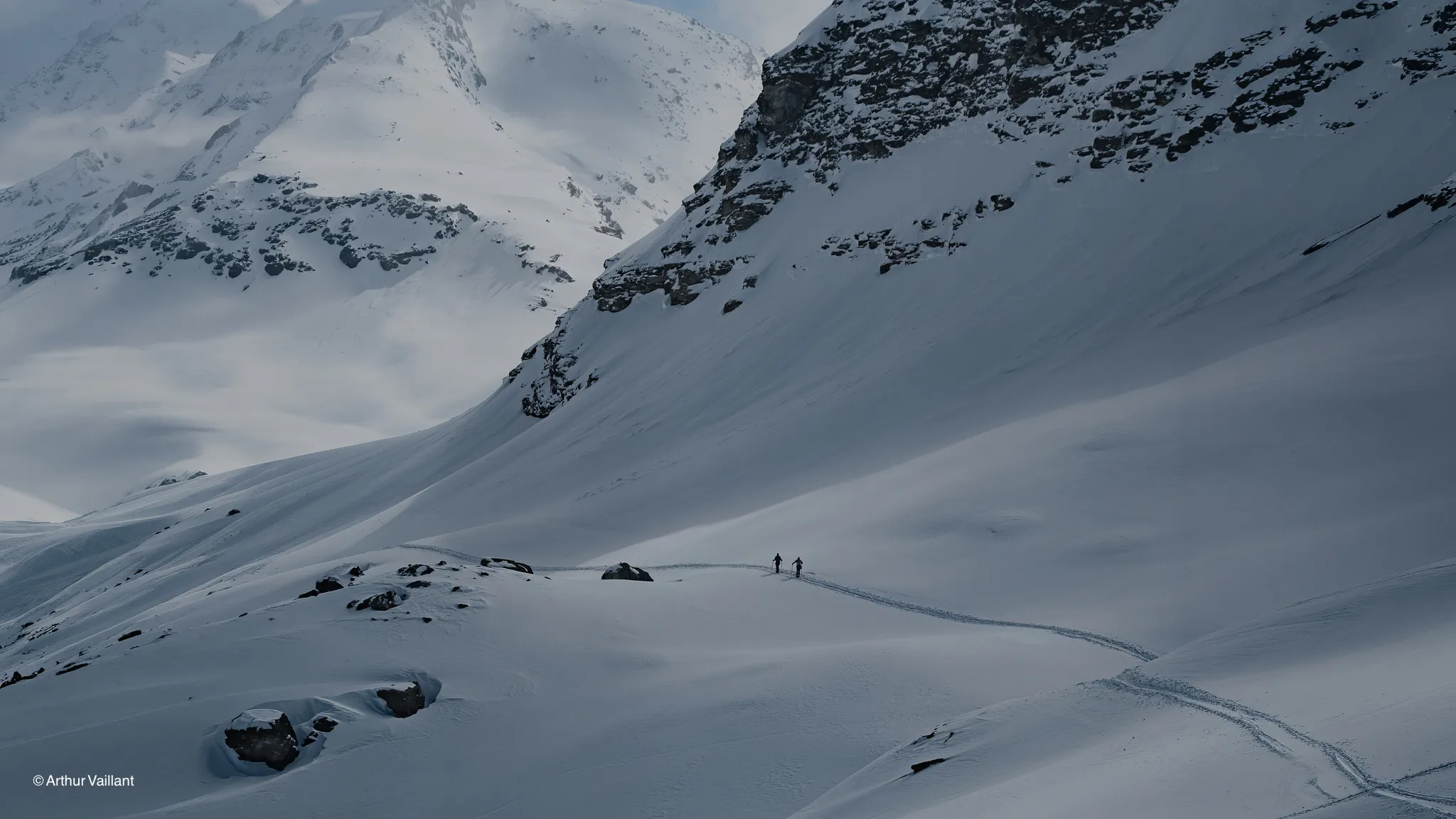Two skiers are making their way across a vast snowfield, surrounded by majestic Alpine mountains.