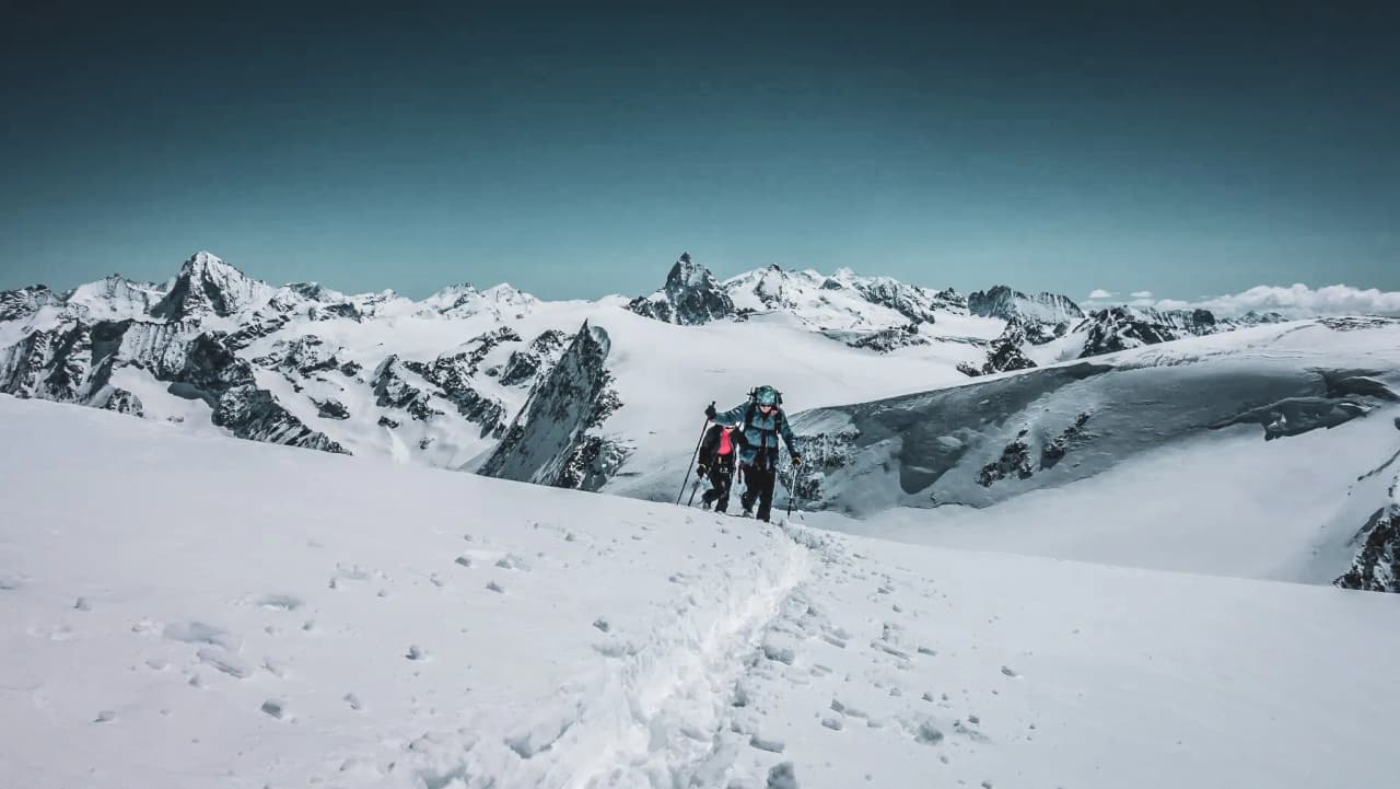 Een hoog berglandschap bedekt met sneeuw, met mountainbikers die zich een weg banen over een pad dat door de sneeuw is gesneden. De lucht is helder, een zachte tint blauw, en de majestueuze pieken van de