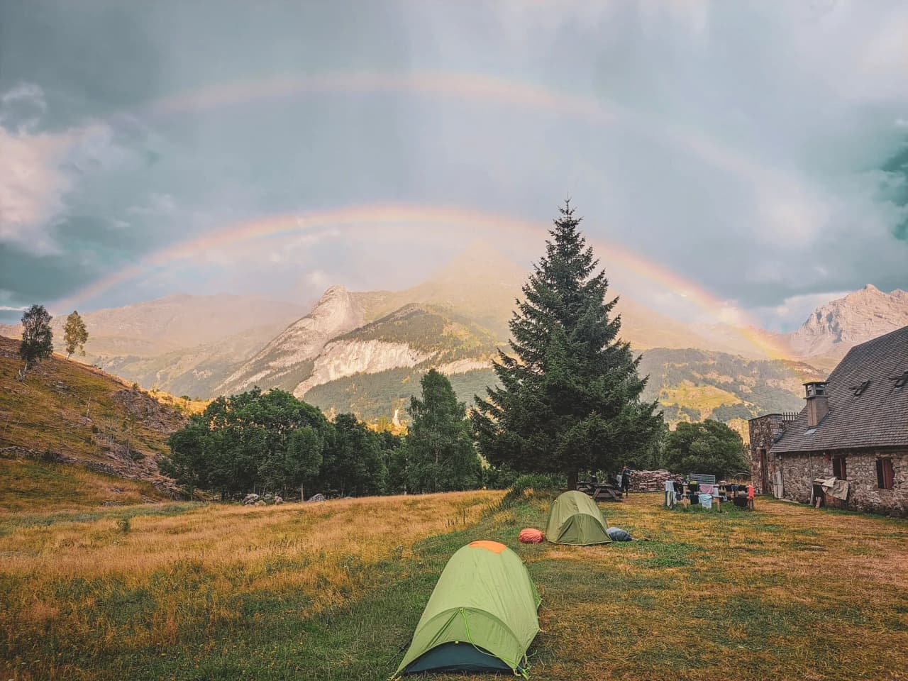 Groene tenten op een zonnige plek, met bergen en regenbogen op de achtergrond.