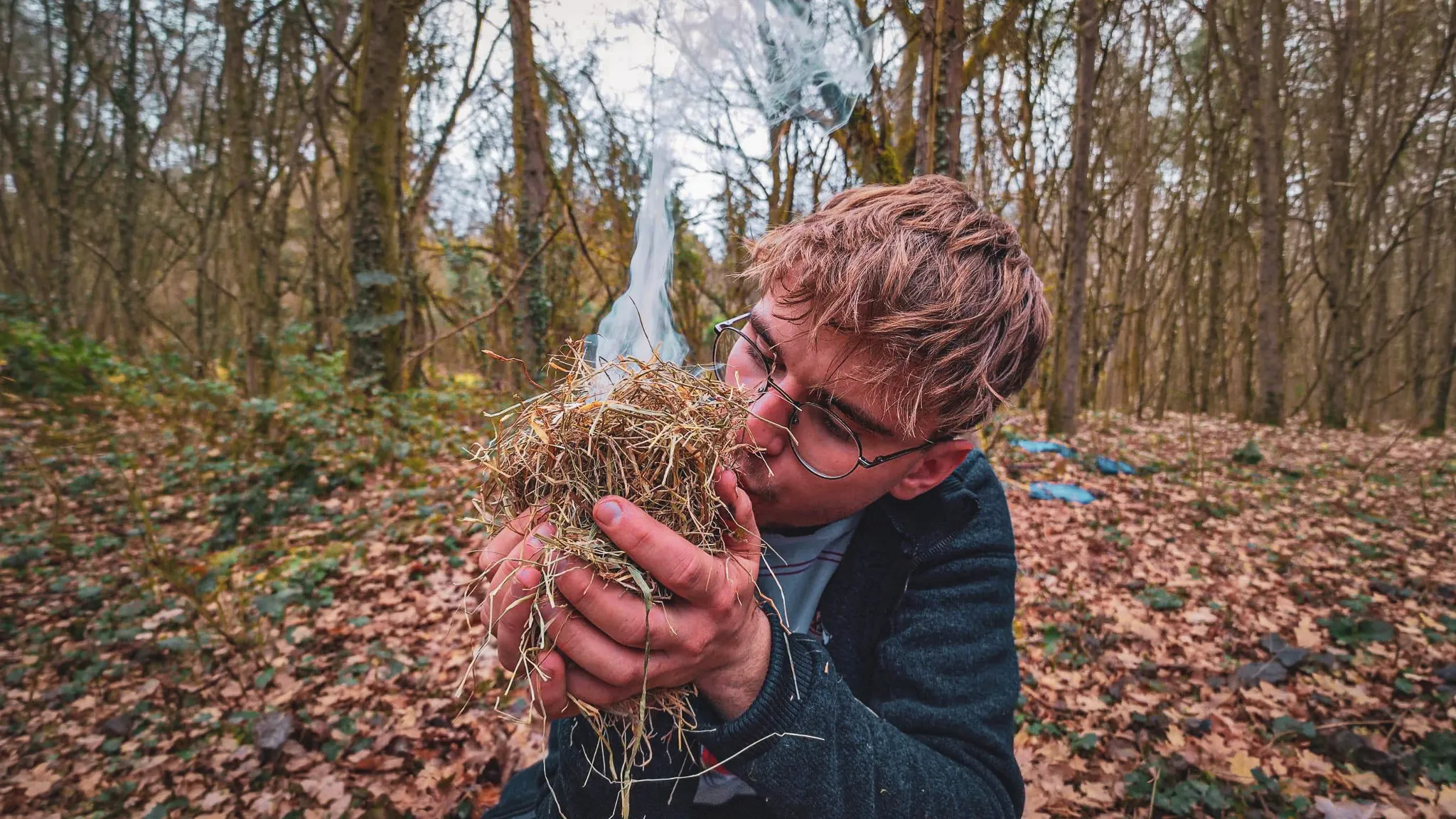 Un participant souffle sur un nid d'herbes dans une forêt, créant une atmosphère d'aventure.