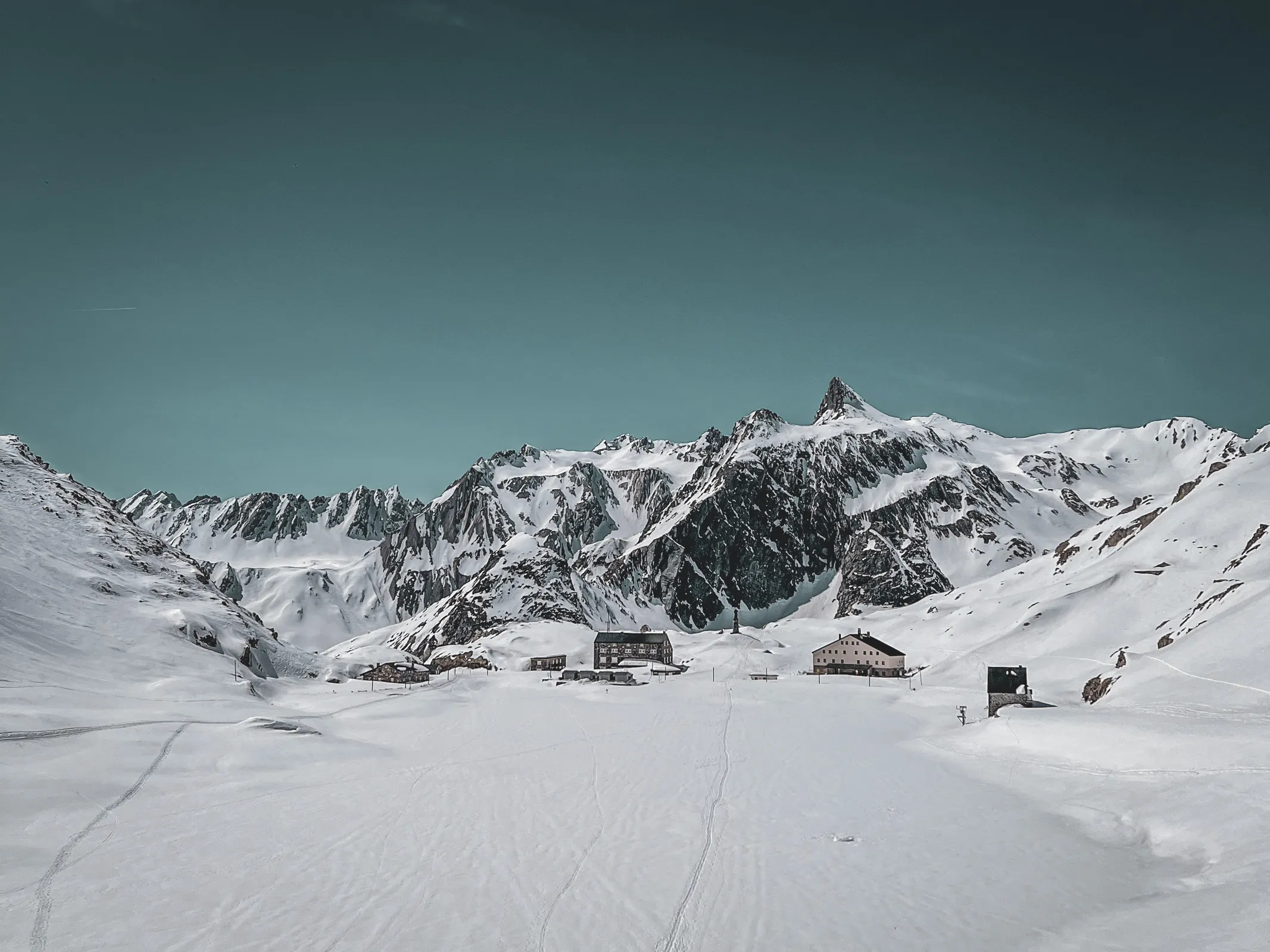 Un paysage alpin enneigé avec des refuges, bordé de sommets majestueux sous un ciel bleu.