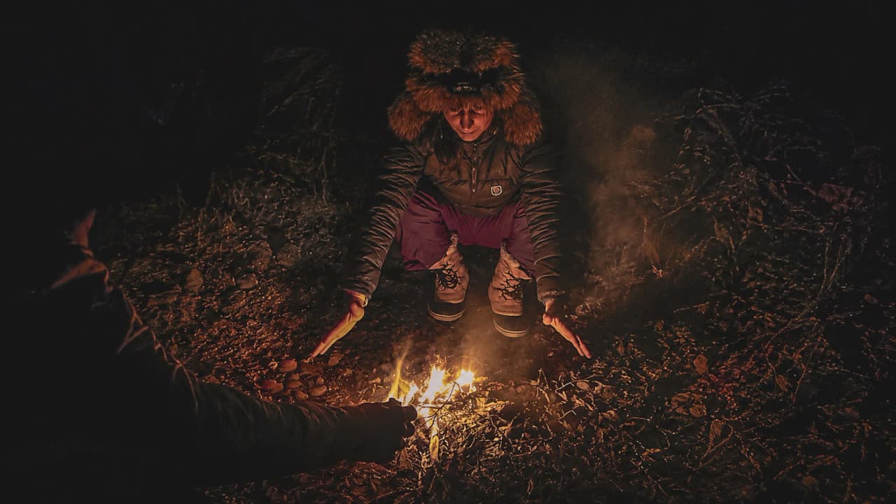 Iemand warmt zijn handen op bij een kampvuur, omringd door de nachtelijke natuur.