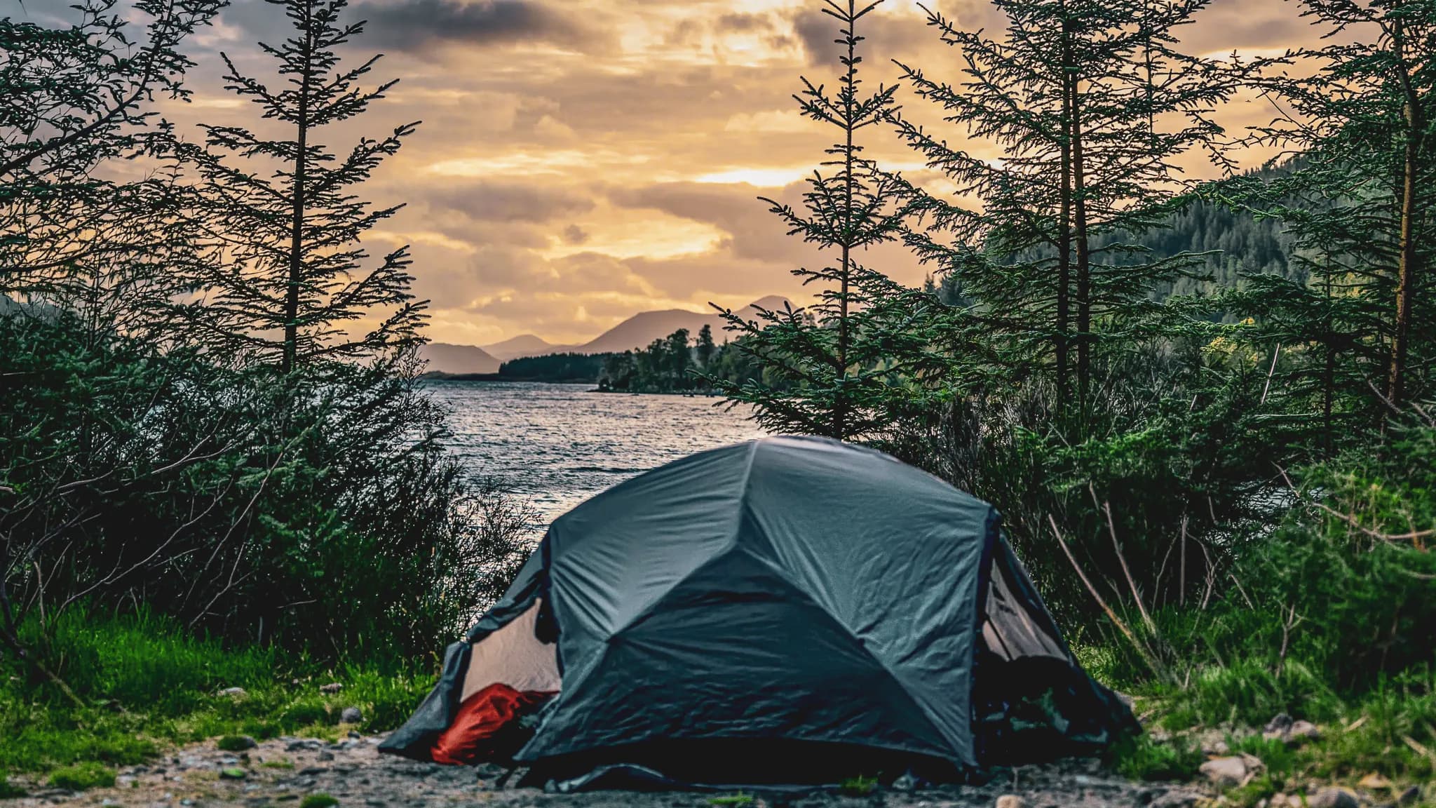 Tent aan de rand van een loch, bergen op de achtergrond onder een gouden hemel, een uitnodiging voor Schots avontuur.