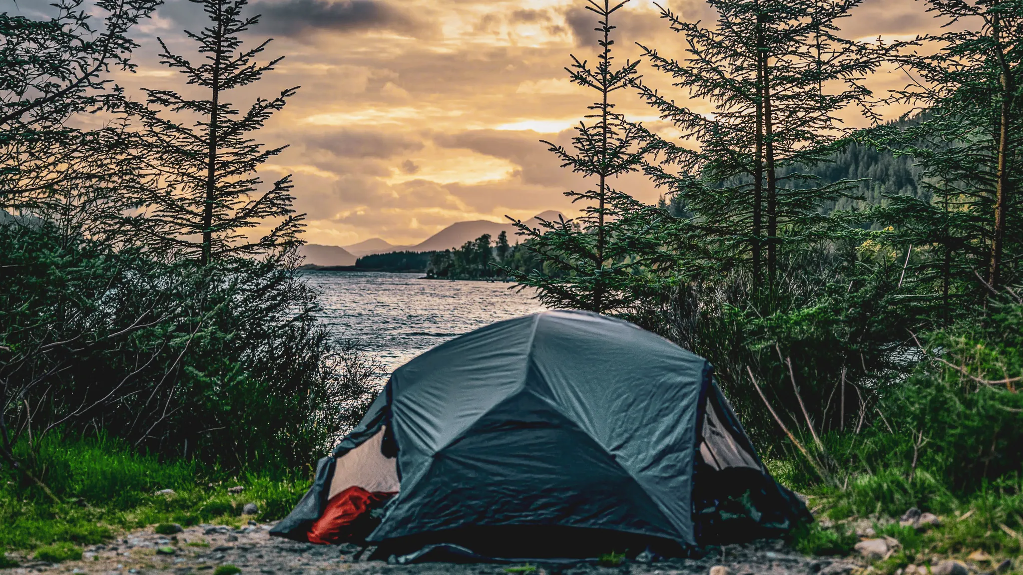 Tent aan de rand van een loch, bergen op de achtergrond onder een gouden hemel, een uitnodiging voor Schots avontuur.