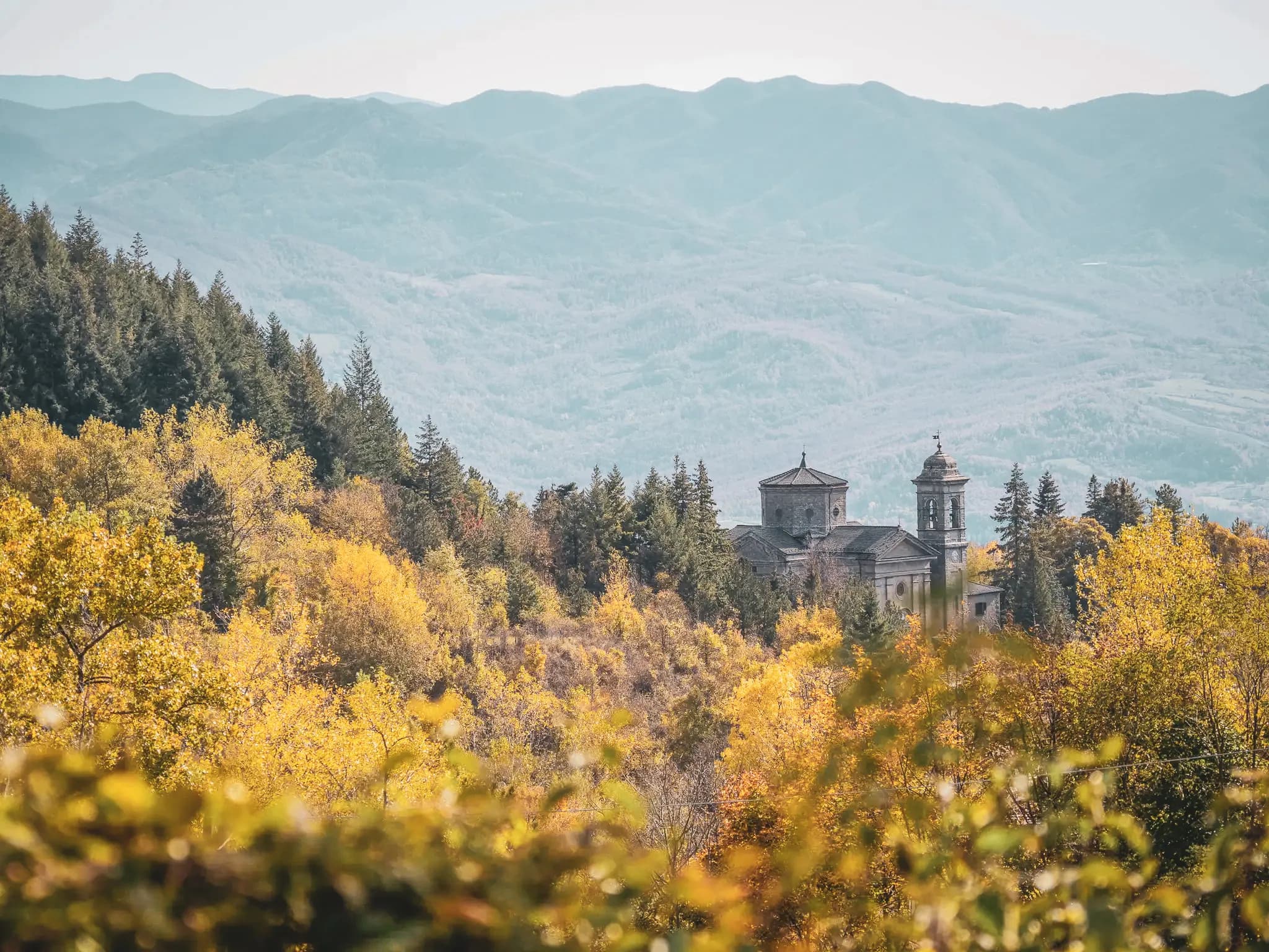 Un paysage automnal avec des arbres aux feuilles jaunes et orange, surplombé par des montagnes en arrière-plan. Au milieu de la scène se trouve un ancien bâtiment de style religieux, probablement une église ou un monastère, flan