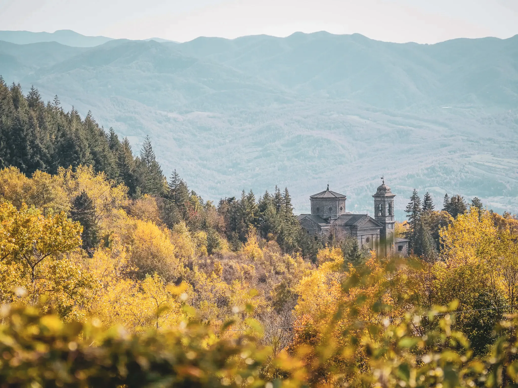 Un paysage automnal avec des arbres aux feuilles jaunes et orange, surplombé par des montagnes en arrière-plan. Au milieu de la scène se trouve un ancien bâtiment de style religieux, probablement une église ou un monastère, flan