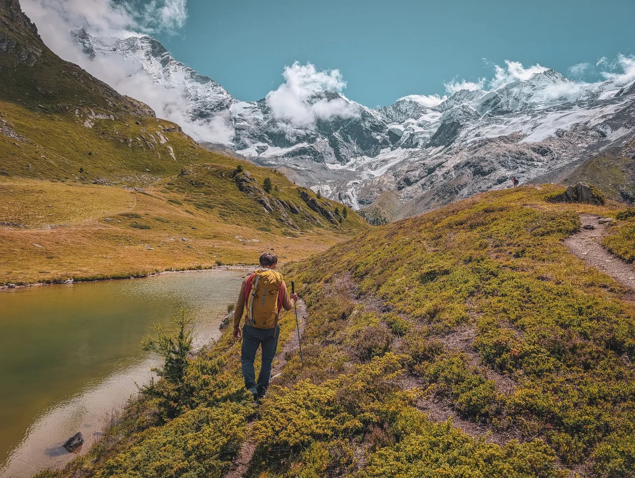 A solitary hiker hugs an Alpine lake, with the majestic snow-capped peaks in the background.