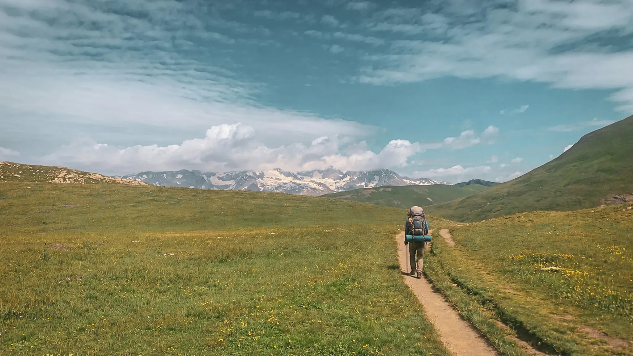 Un randonneur sur un sentier verdoyant, entouré de montagnes majestueuses sous un ciel bleu.