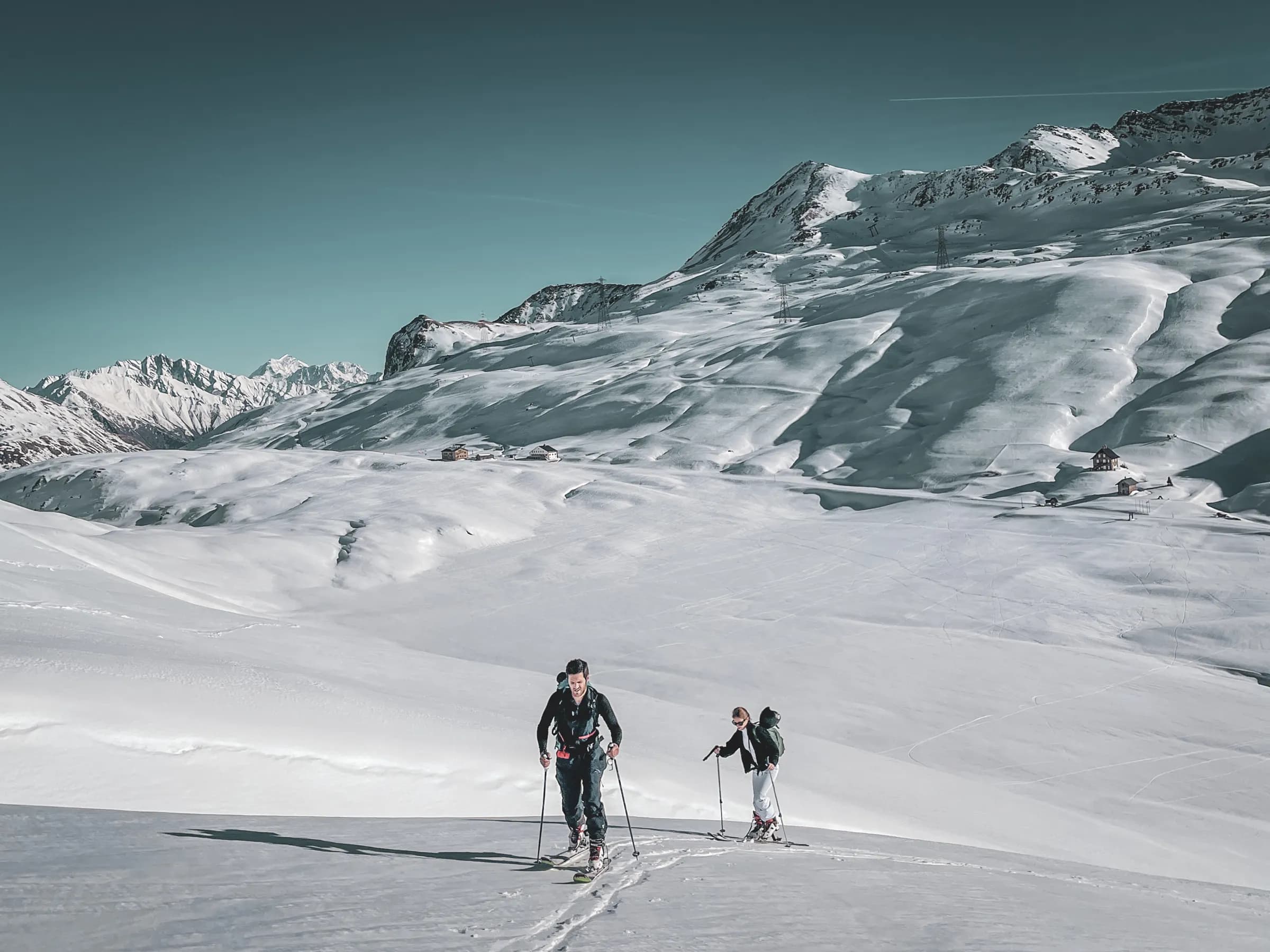Two touring skiers cross a snow-covered alpine landscape, with majestic mountains in the background.