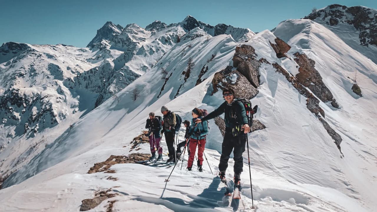 Groupe de skieurs randonneurs sur un sommet enneigé, avec les majestueuses Alpes en arrière-plan.