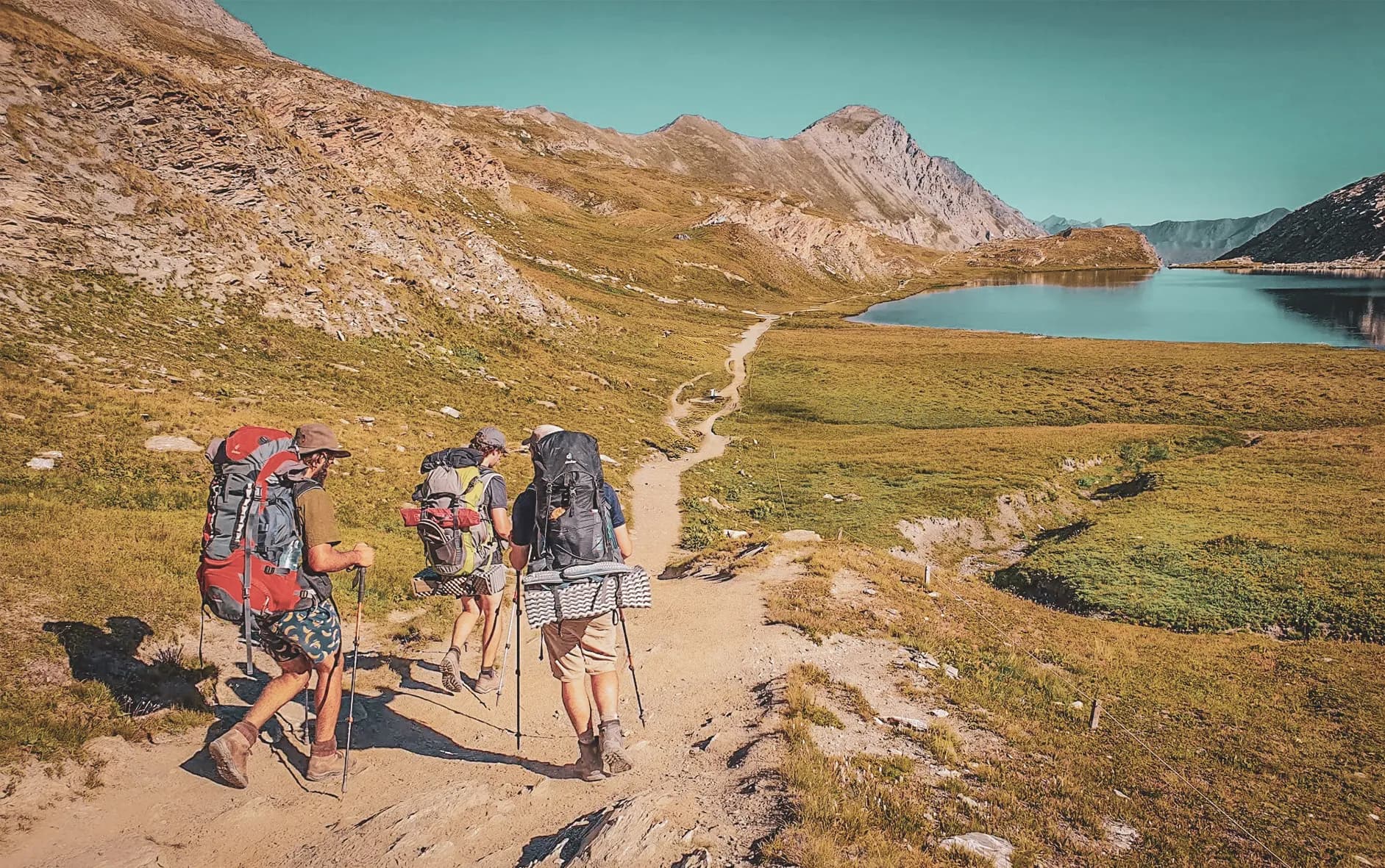 Three hikers walk along a picturesque path beside an Alpine lake, with mountains in the background.
