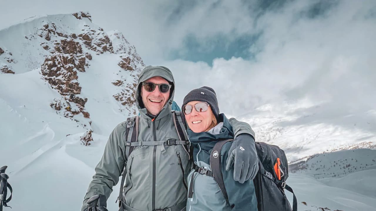 Un couple souriant en ski de randonnée, avec des sommets enneigés en arrière-plan.