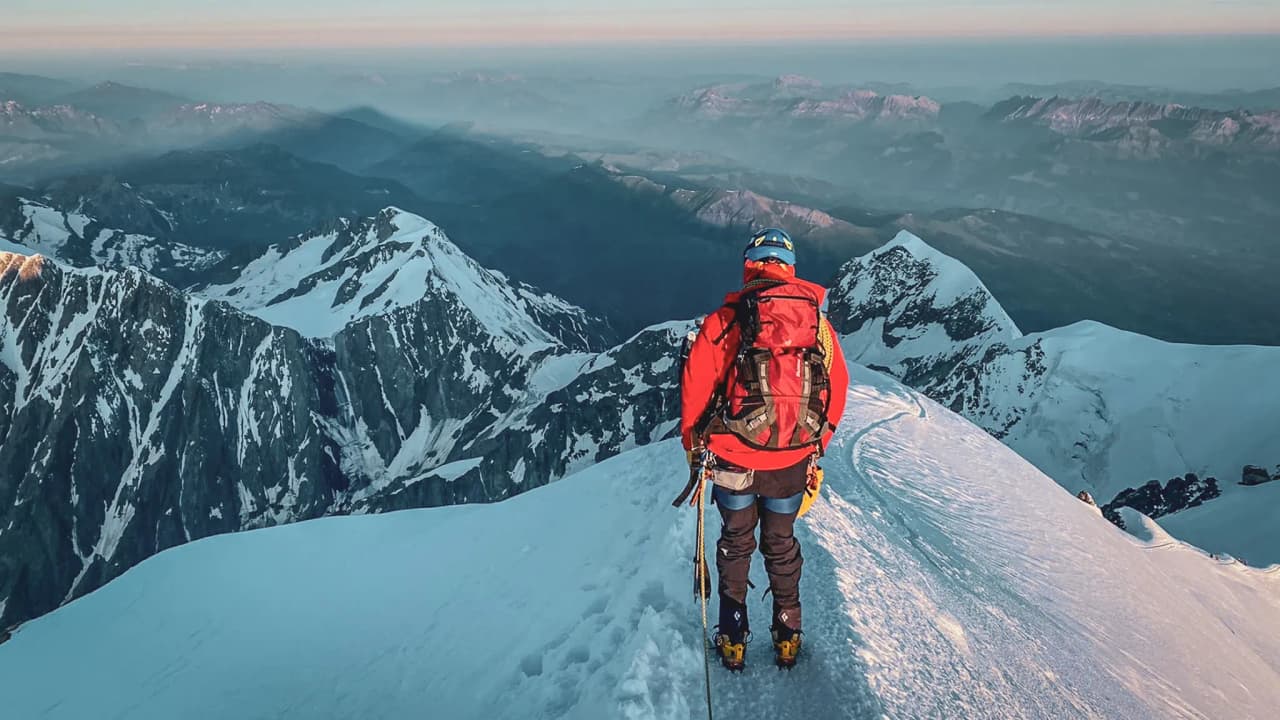 Een bergbeklimmer in een rode jas kijkt naar de besneeuwde toppen van de Mont Blanc bij zonsopgang.