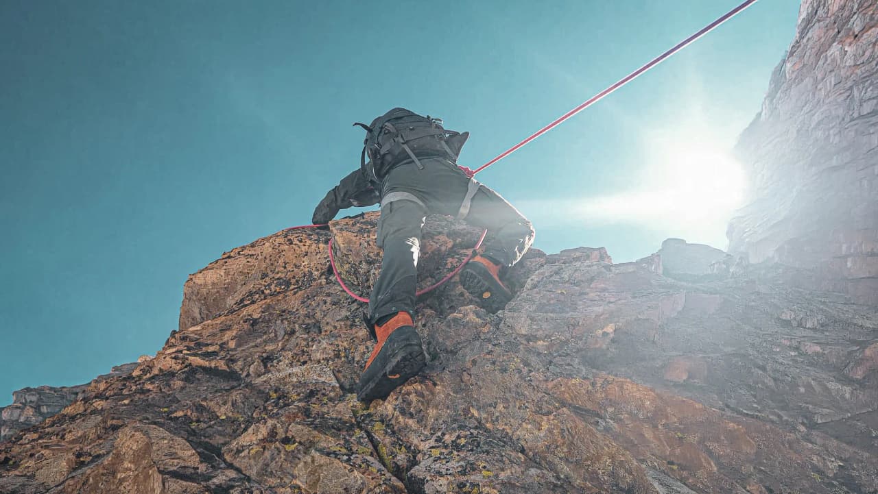 Grimpeur en pleine ascension sur un rocher sous un ciel bleu, lumière éclatante en arrière-plan.