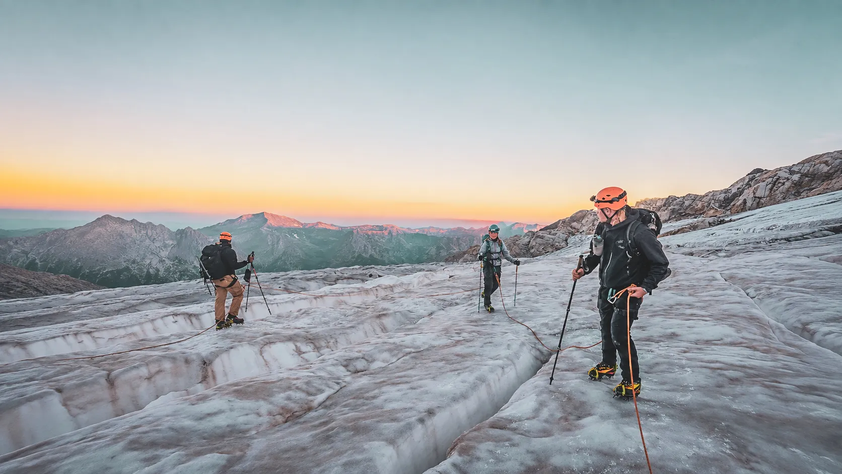 Groupe d'alpinistes sur un glacier au lever du soleil, avec des sommets majestueux en arrière-plan.