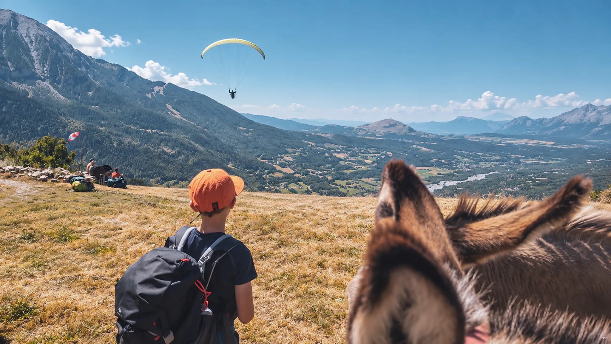 Un enfant avec un âne admire un parapentiste surplombant les montagnes des Écrins.