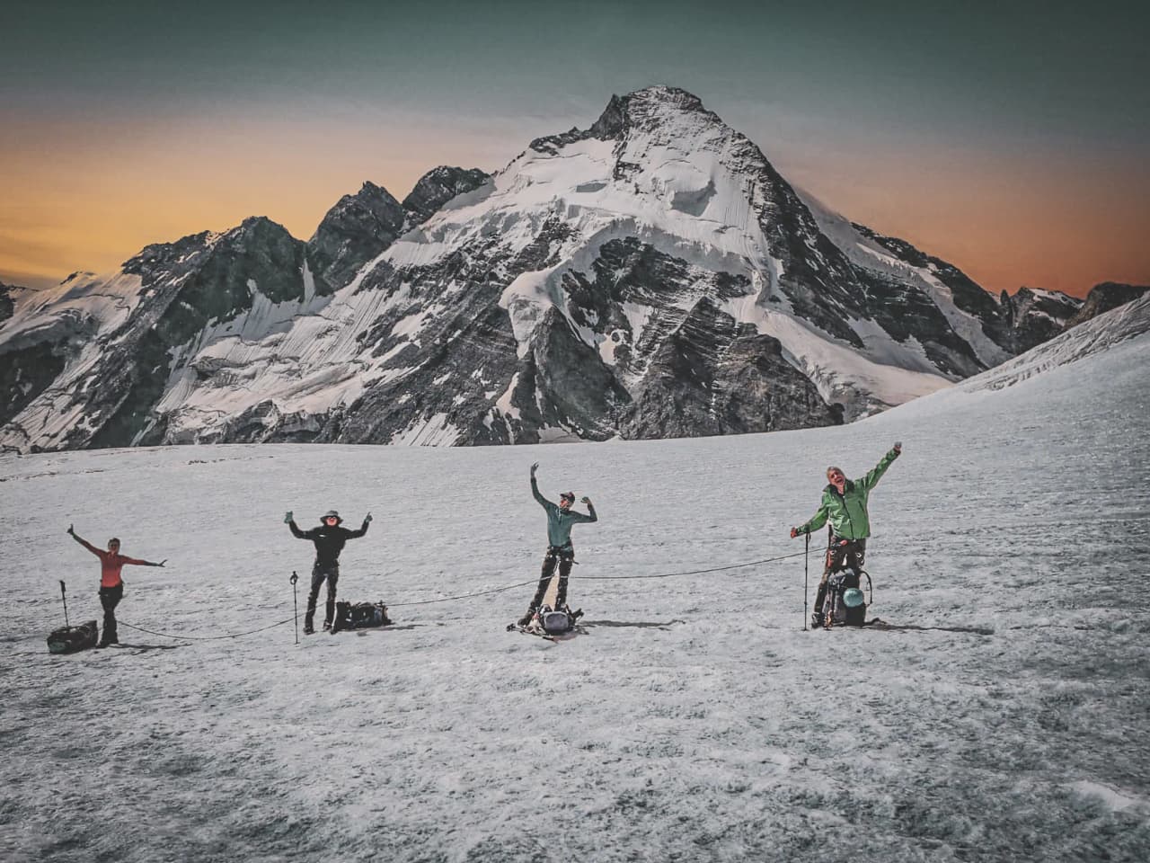 Four climbers on a glacier, raising their arms with majestic peaks in the background.