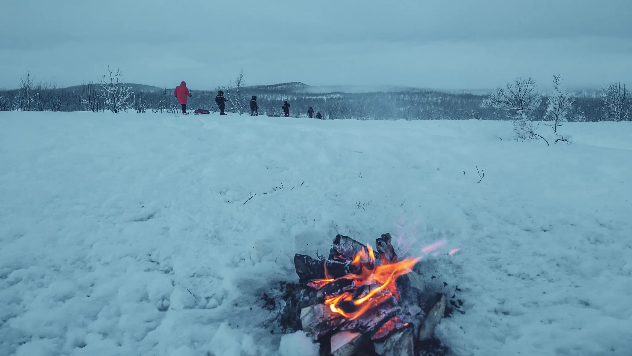 Een gezellig kampvuur in een besneeuwd landschap, met avonturiers op sneeuwschoenen aan de horizon.