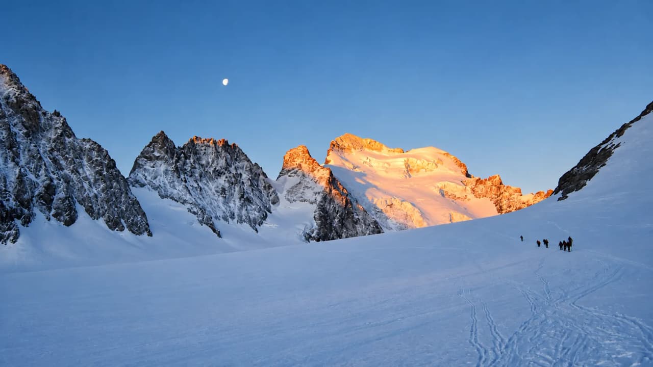Een touwgroep op de gletsjer bij dageraad, met de toppen van de Écrins in het eerste zonlicht en de maan nog zichtbaar.