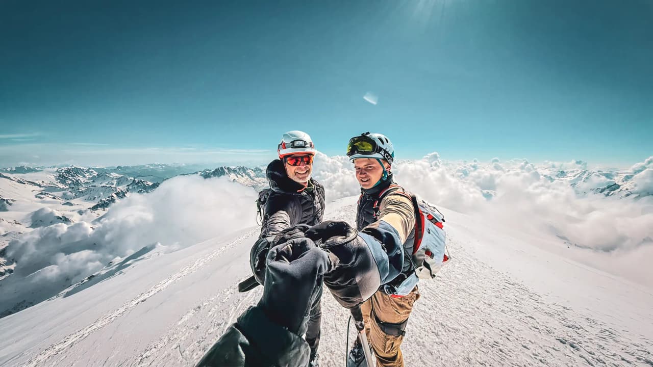 Two smiling mountaineers on the summit of Mont Blanc, surrounded by clouds and majestic peaks.