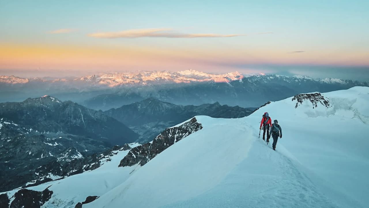 Deux alpinistes marchent sur un glacier, face à un panorama époustouflant au lever du soleil.