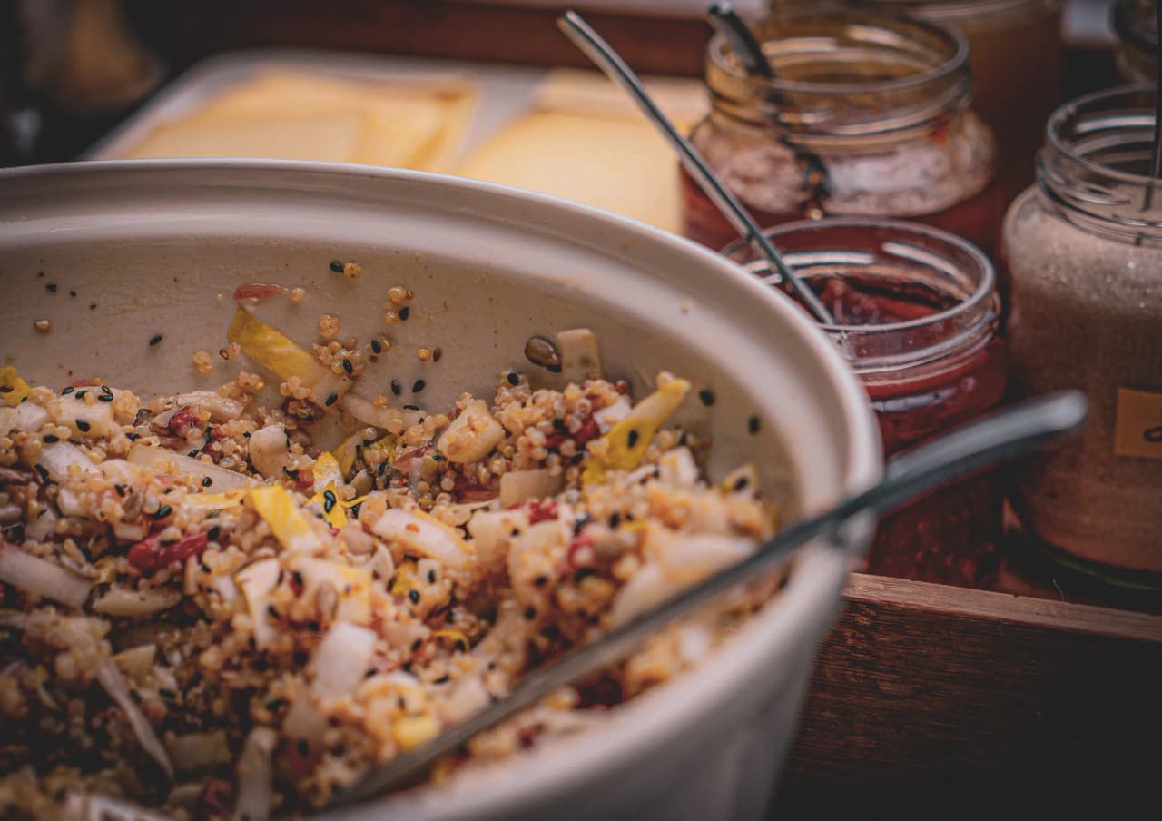 Een schaal met quinoa is gevuld met verschillende groenten, zoals stukjes gele paprika en ui, en zaden. Op de achtergrond staan een aantal glazen potten met verschillende kruiden en ingrediënten, zoals
