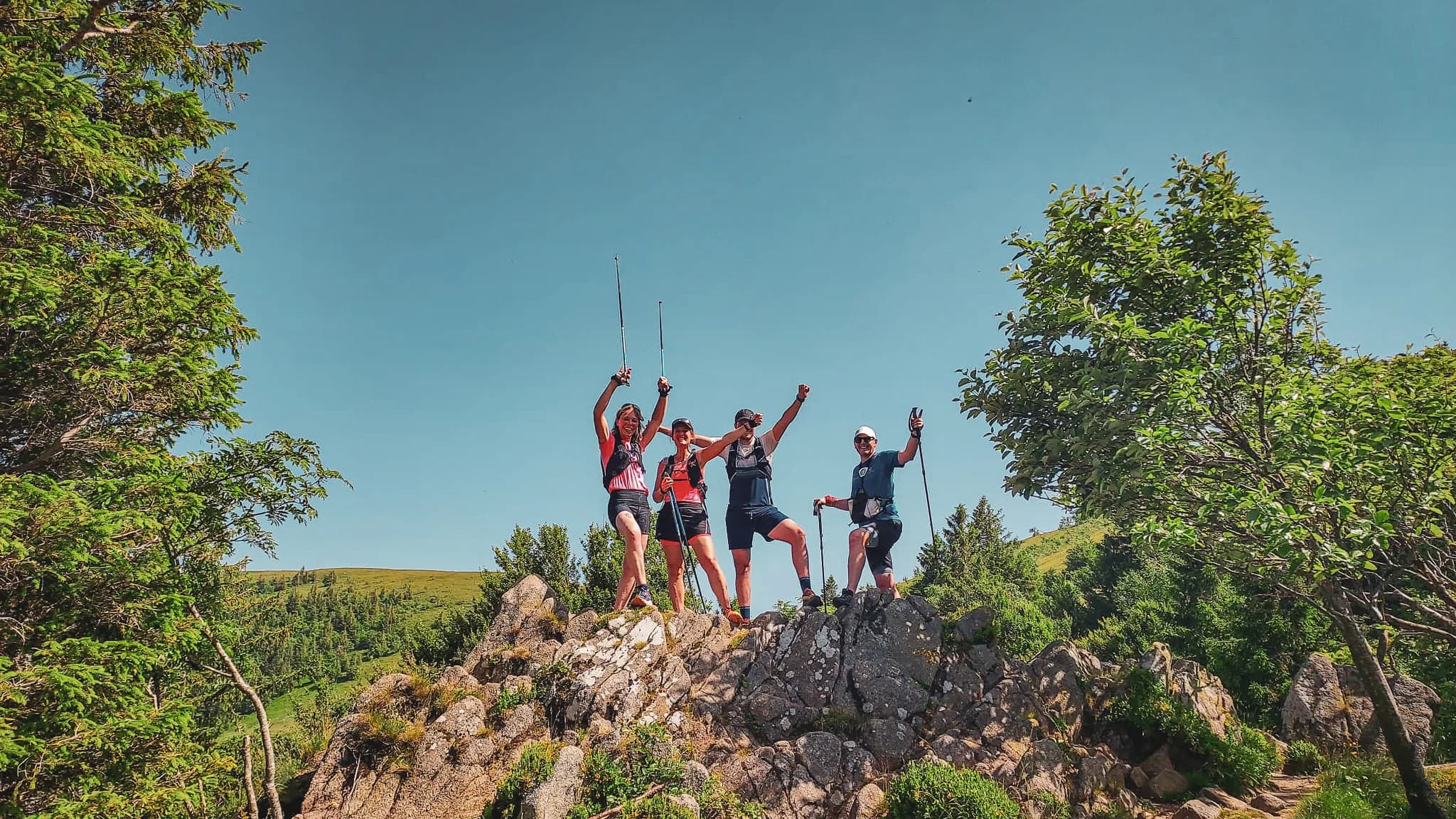 Quatre randonneurs jubilent sur un rocher avec vue sur les Vosges, sous un ciel bleu éclatant.