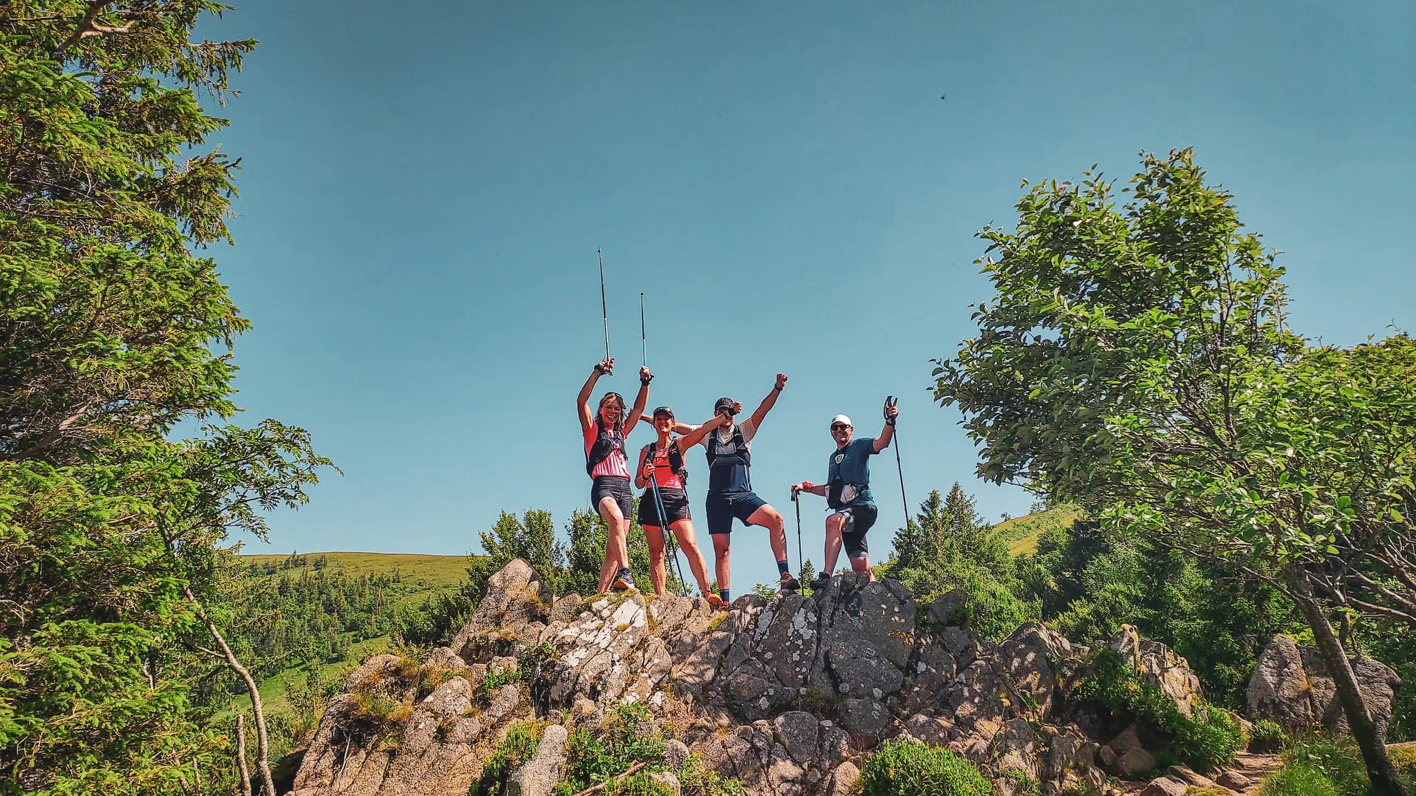 Quatre randonneurs jubilent sur un rocher avec vue sur les Vosges, sous un ciel bleu éclatant.