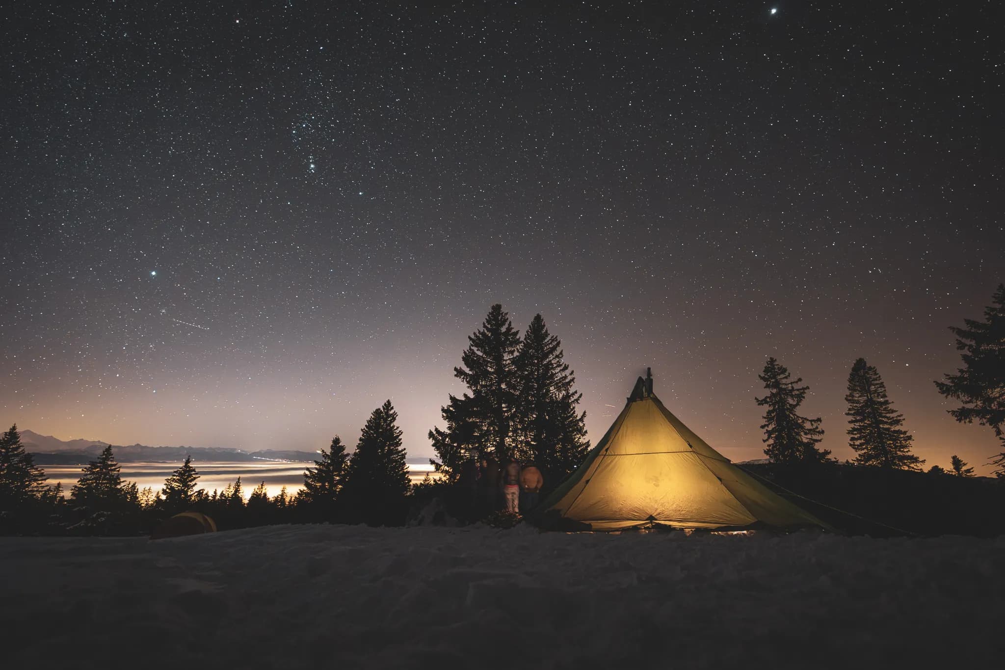 Under a starry sky, a luminous tent stands in the snow, surrounded by majestic fir trees.