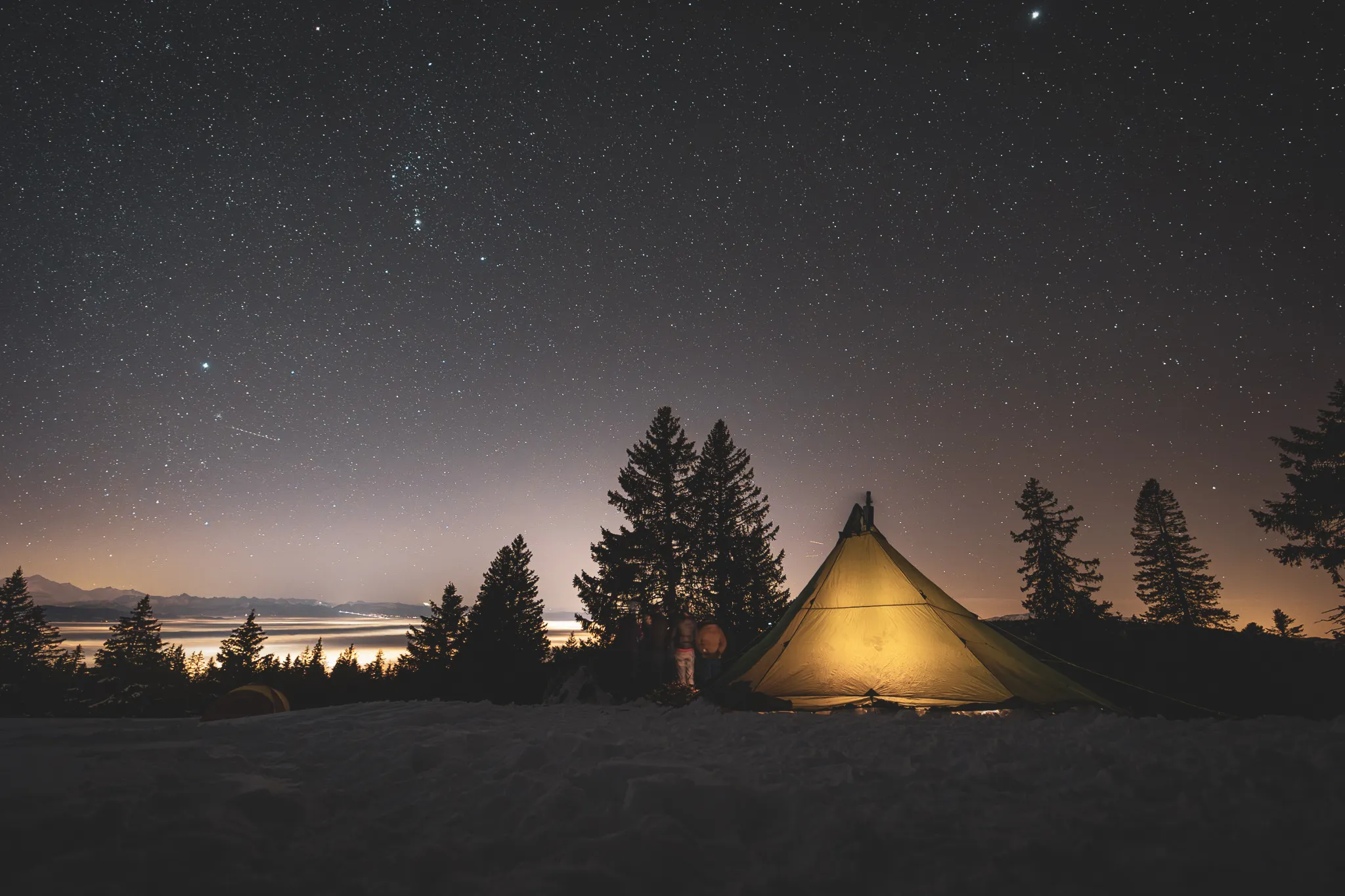 Under a starry sky, a luminous tent stands in the snow, surrounded by majestic fir trees.