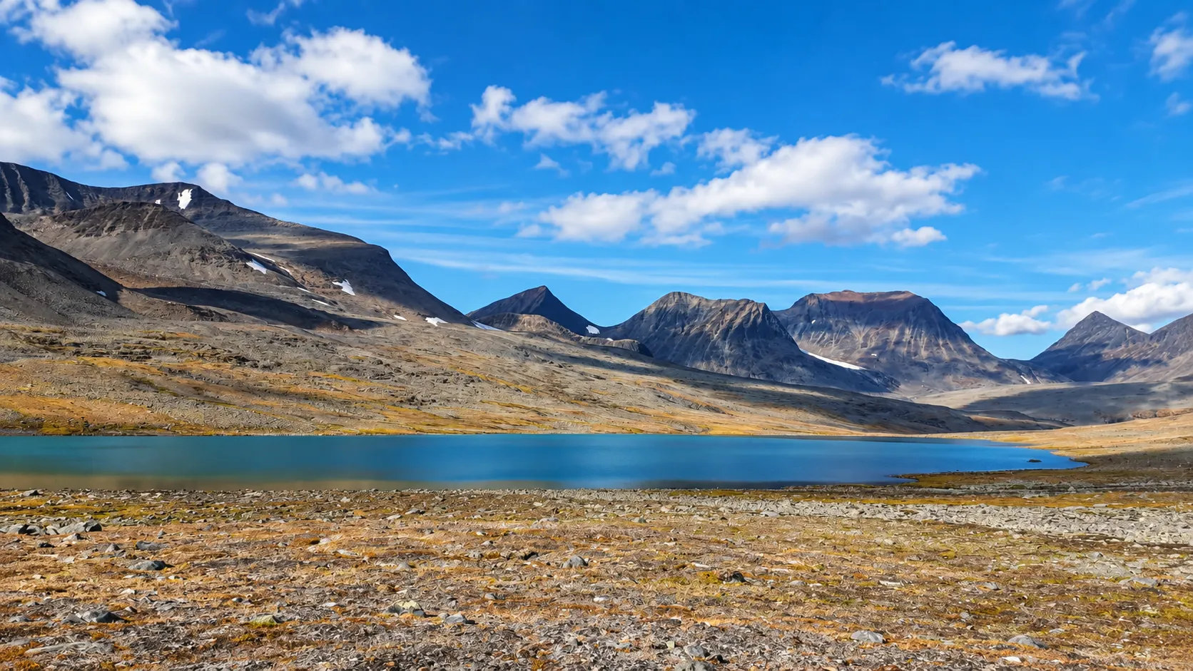 Een majestueus berglandschap in Lapland, met een turkoois meer onder een blauwe hemel bezaaid met wolken.