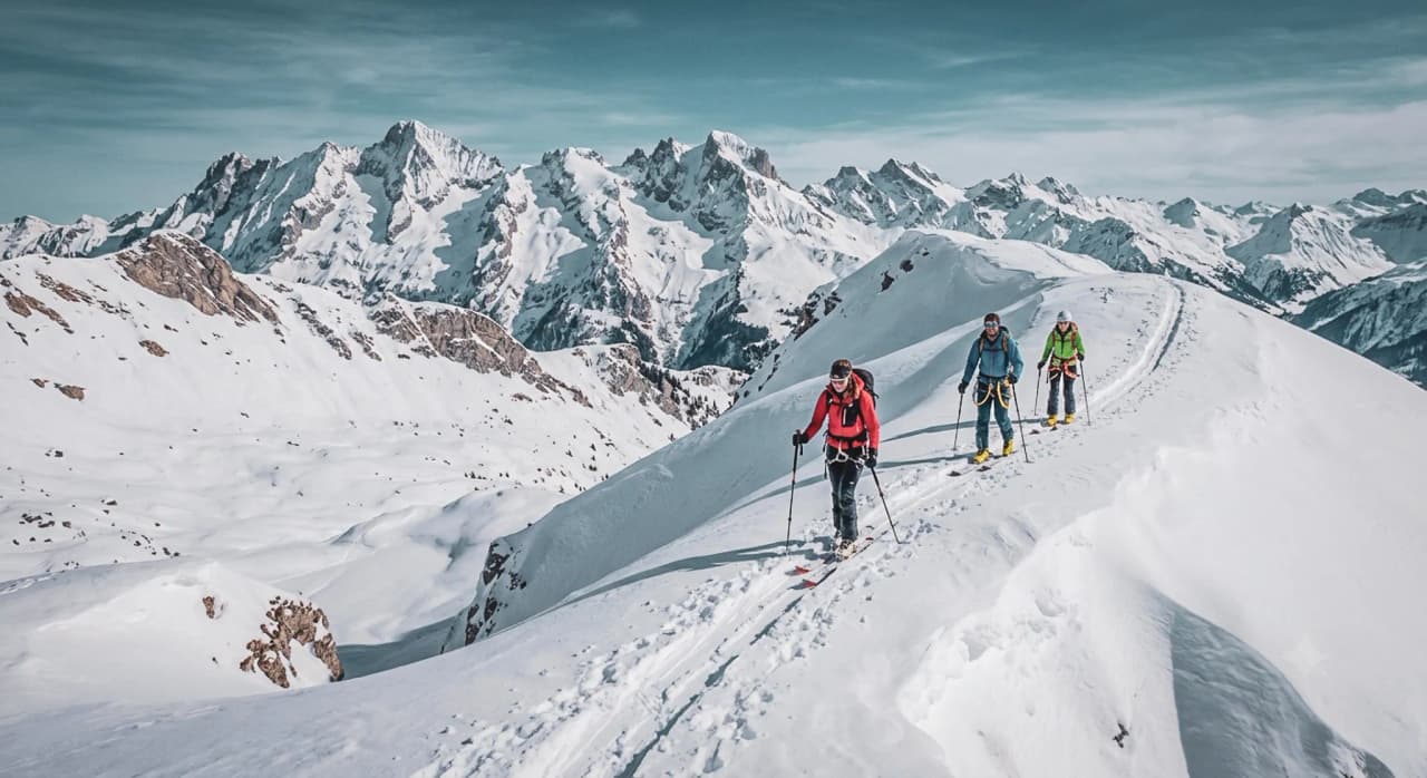 A group of ski tourers on a snow-covered ridge, with the majestic Alps in the background.