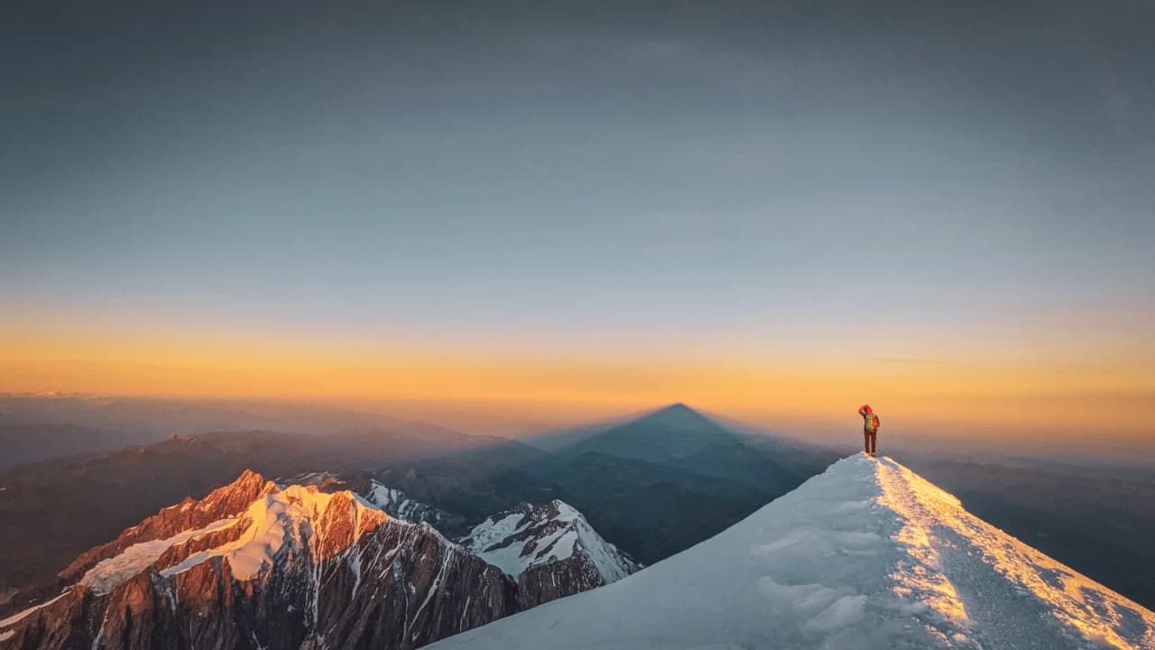Un alpiniste sur un sommet enneigé, face à un coucher de soleil époustouflant en montagne.