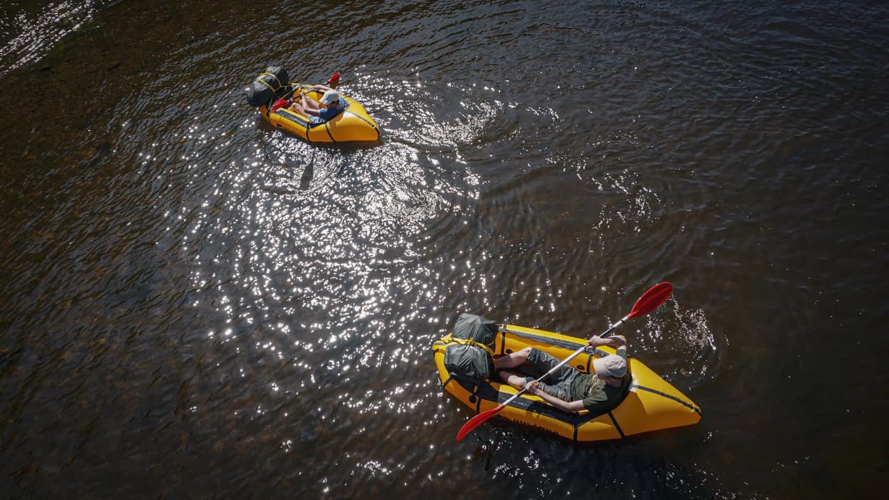 Twee gele packrafts glijden over een glinsterende rivier, omringd door weelderige natuur.