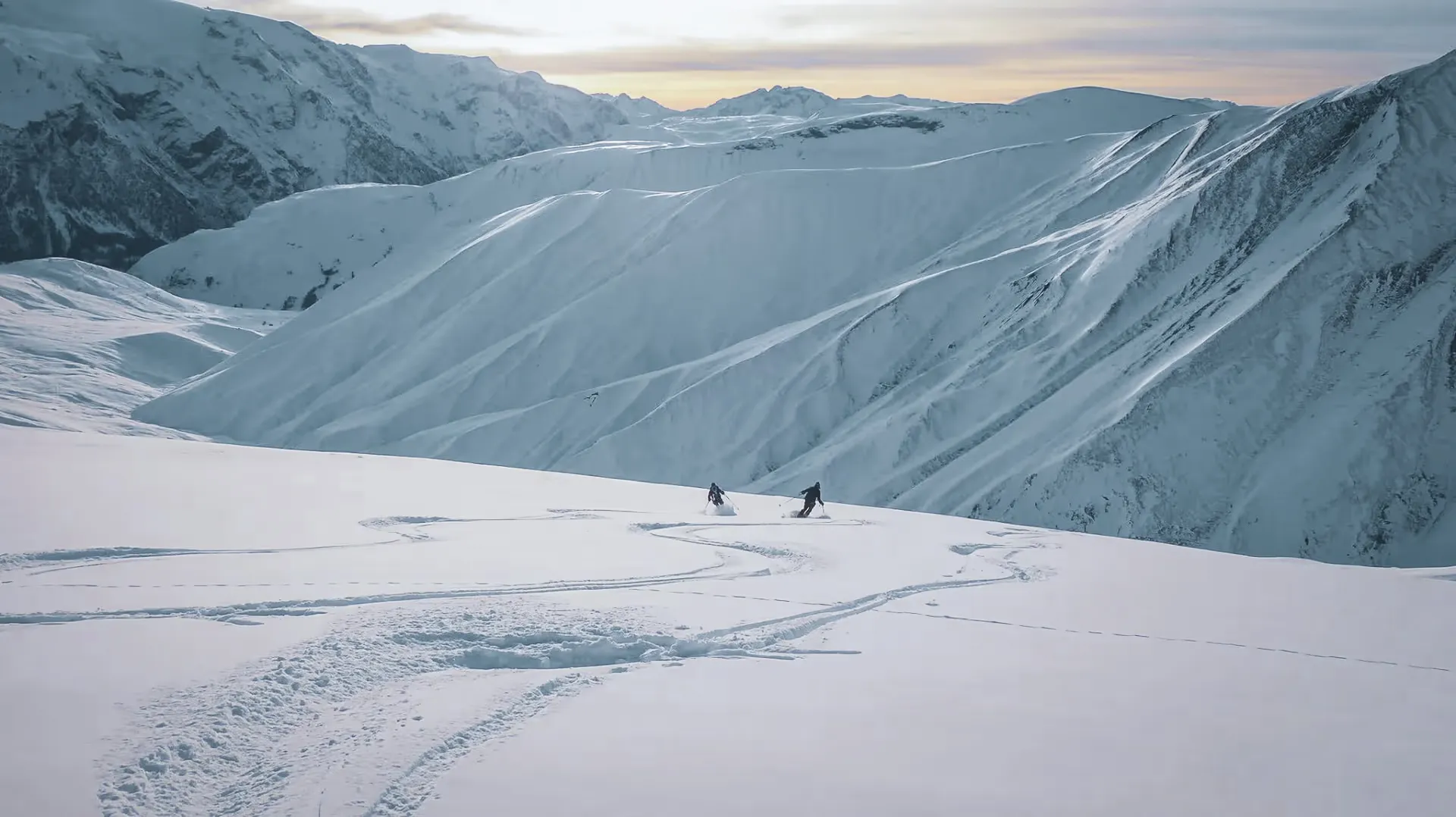 Two skiers set off down snowy slopes, illuminated by a gentle sunset.