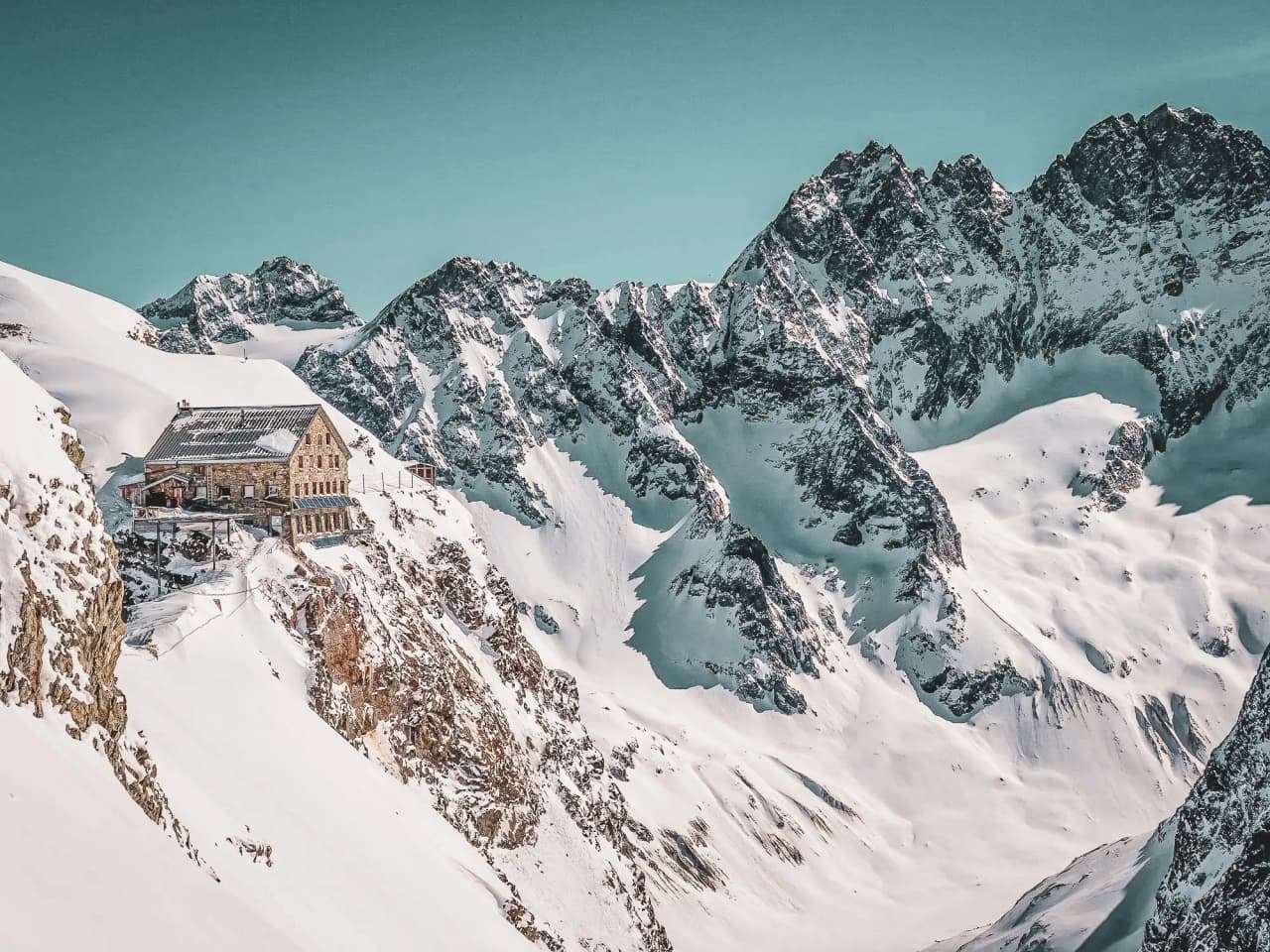 Een stenen berghut op een besneeuwde top, omgeven door berglandschap. Een trap leidt naar de hut, die een metalen luifel heeft. Een skiër met een rugzak