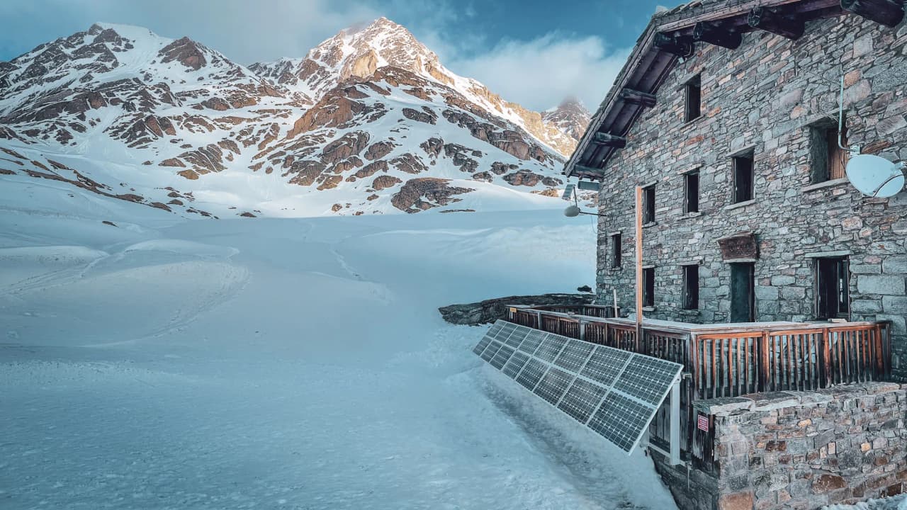 A stone Mountain hut set in an alpine landscape, surrounded by snow and majestic mountains.