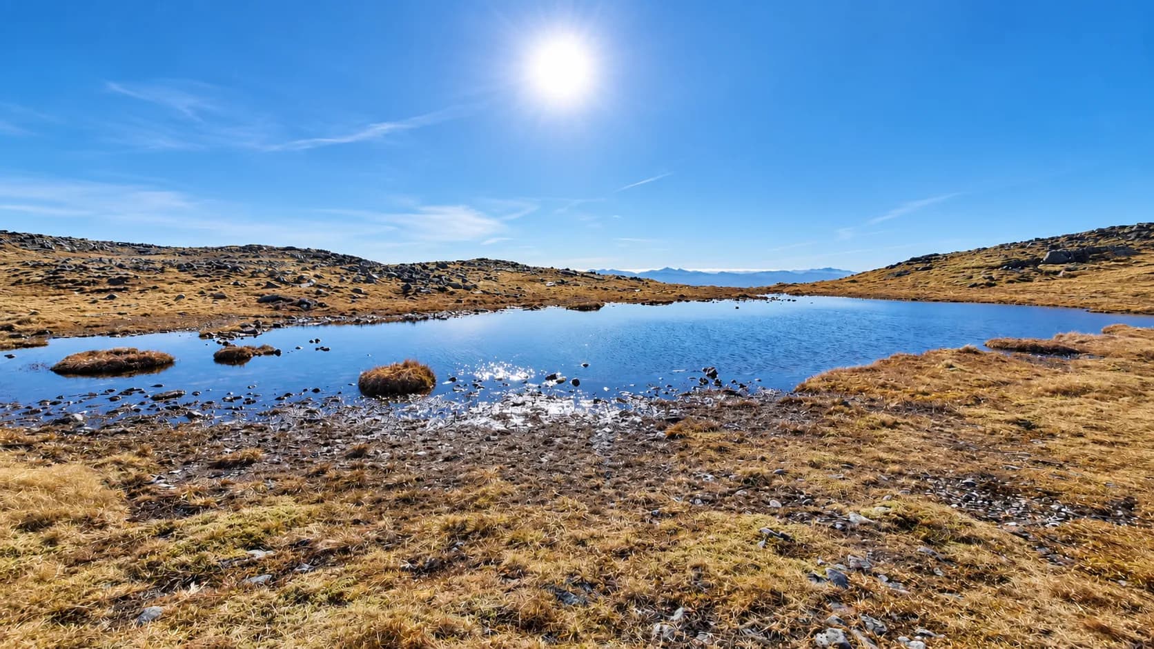 Een klein hooggelegen meertje dat glinstert in de zon, omringd door goudkleurige heide op een plateau in de Highlands