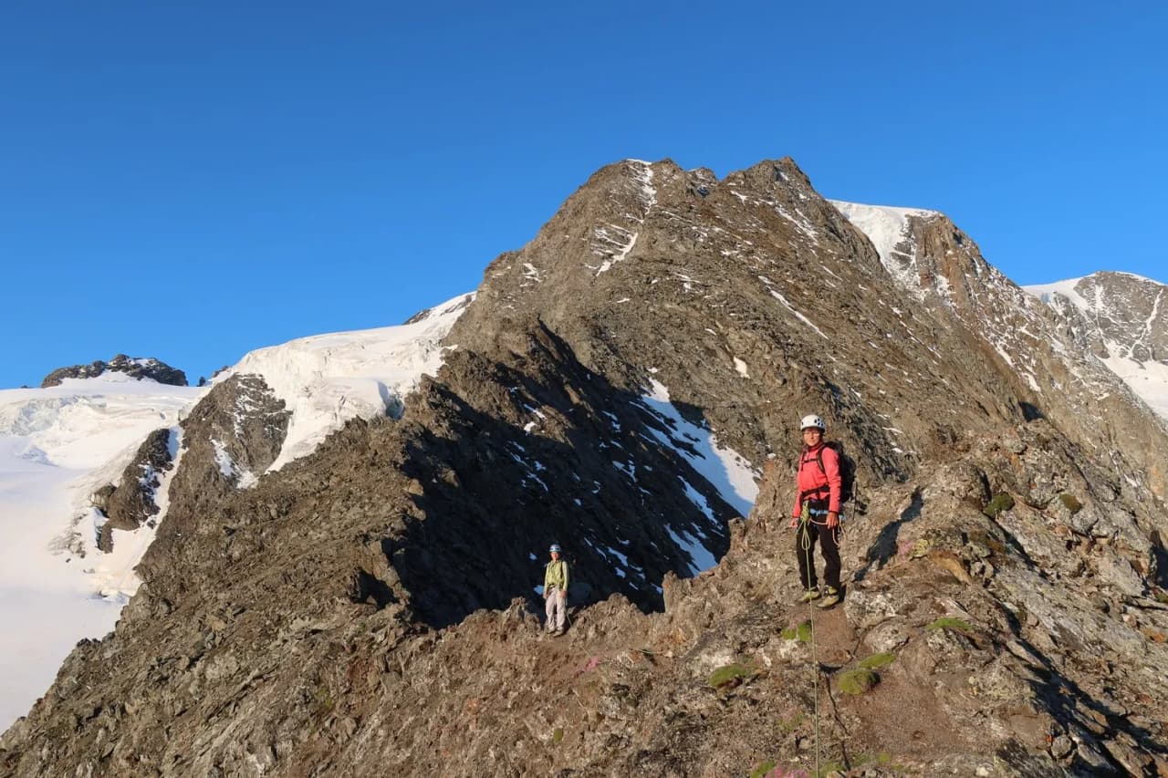 Two climbers on a rocky ridge, with Mont Blanc in the background under a brilliant blue sky.