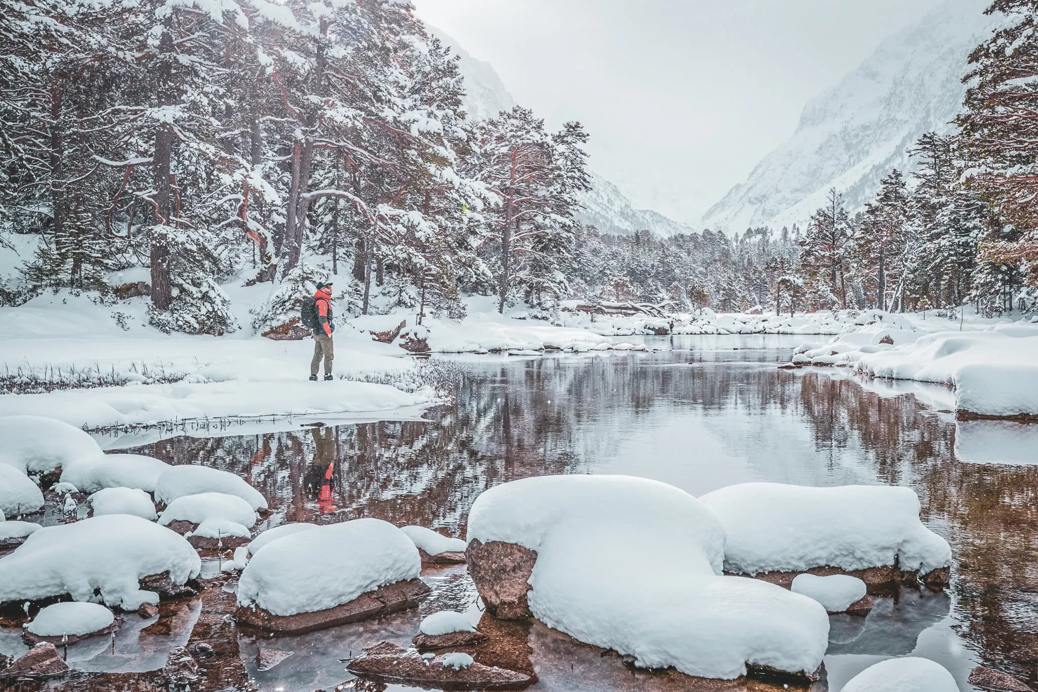 A snowshoe hiker admires the snowy landscape around a peaceful lake in the Pyrenees.