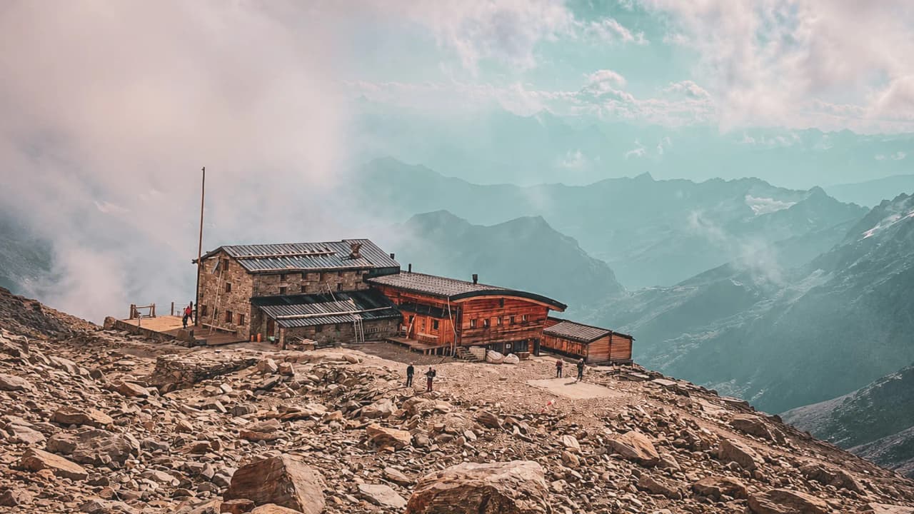 A picturesque mountain hut beneath a blue sky, surrounded by majestic mountains and mysterious clouds.