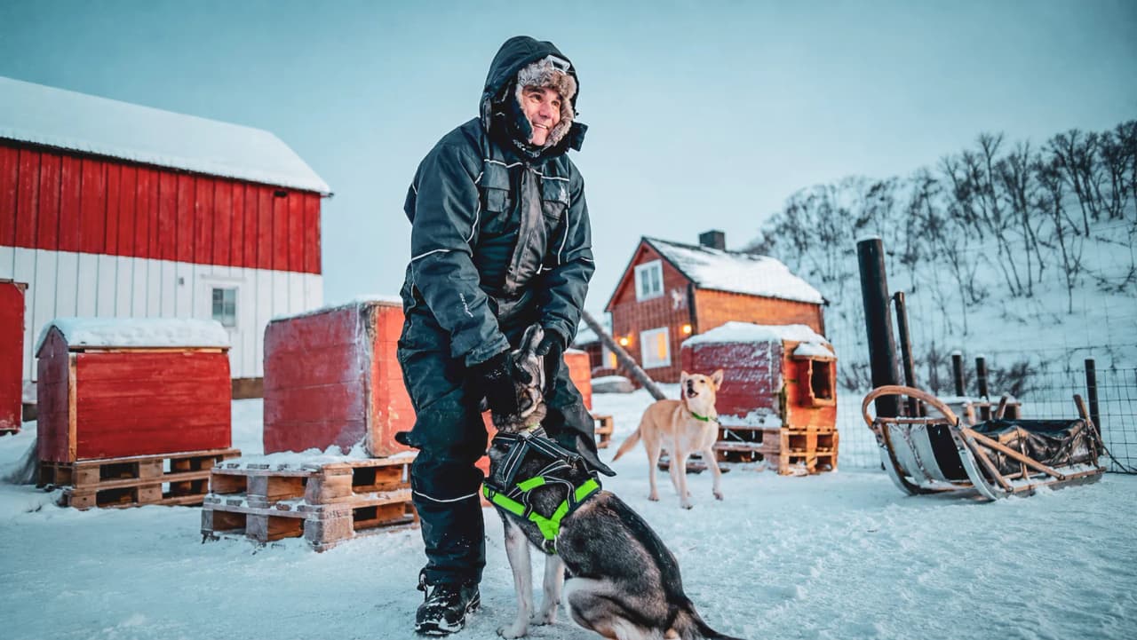 Un musher en combinaison noire caresse un husky dans un paysage enneigé pittoresque.
