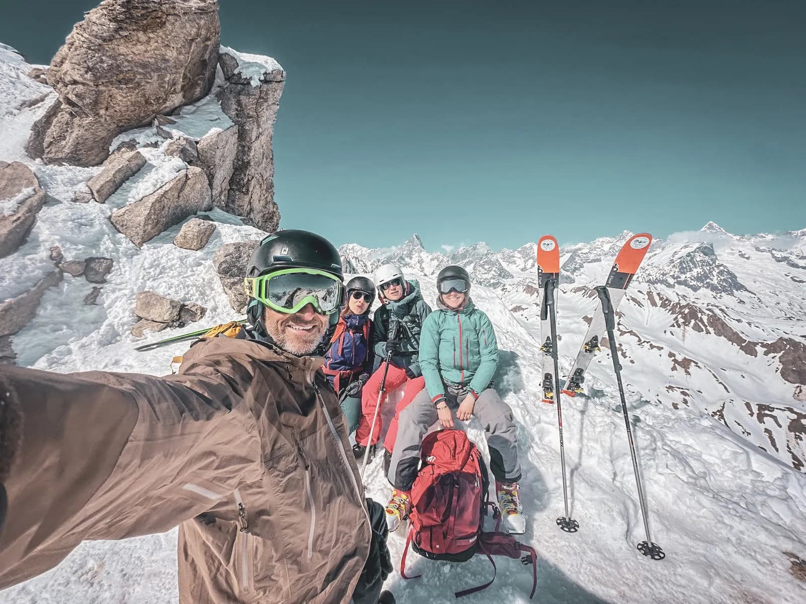 Quatre amis profitent du soleil sur un sommet alpin, ski en main, avec des paysages majestueux en arrière-plan.