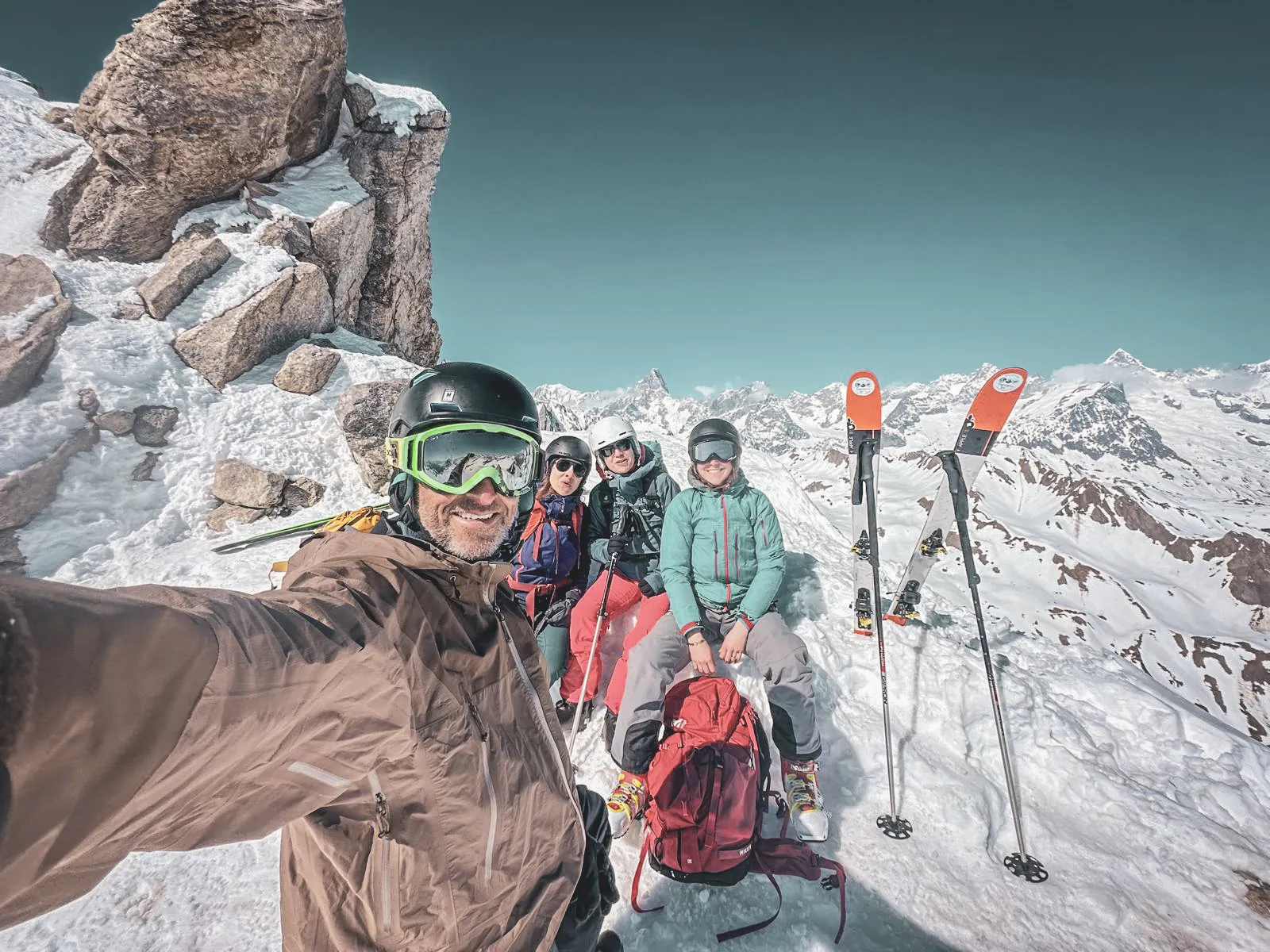 Quatre amis profitent du soleil sur un sommet alpin, ski en main, avec des paysages majestueux en arrière-plan.